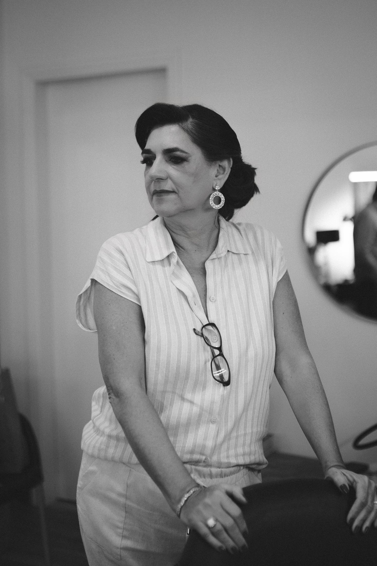Black and white photo of an elegant middle-aged woman wearing striped blouse, large earrings, and glasses hanging from collar.