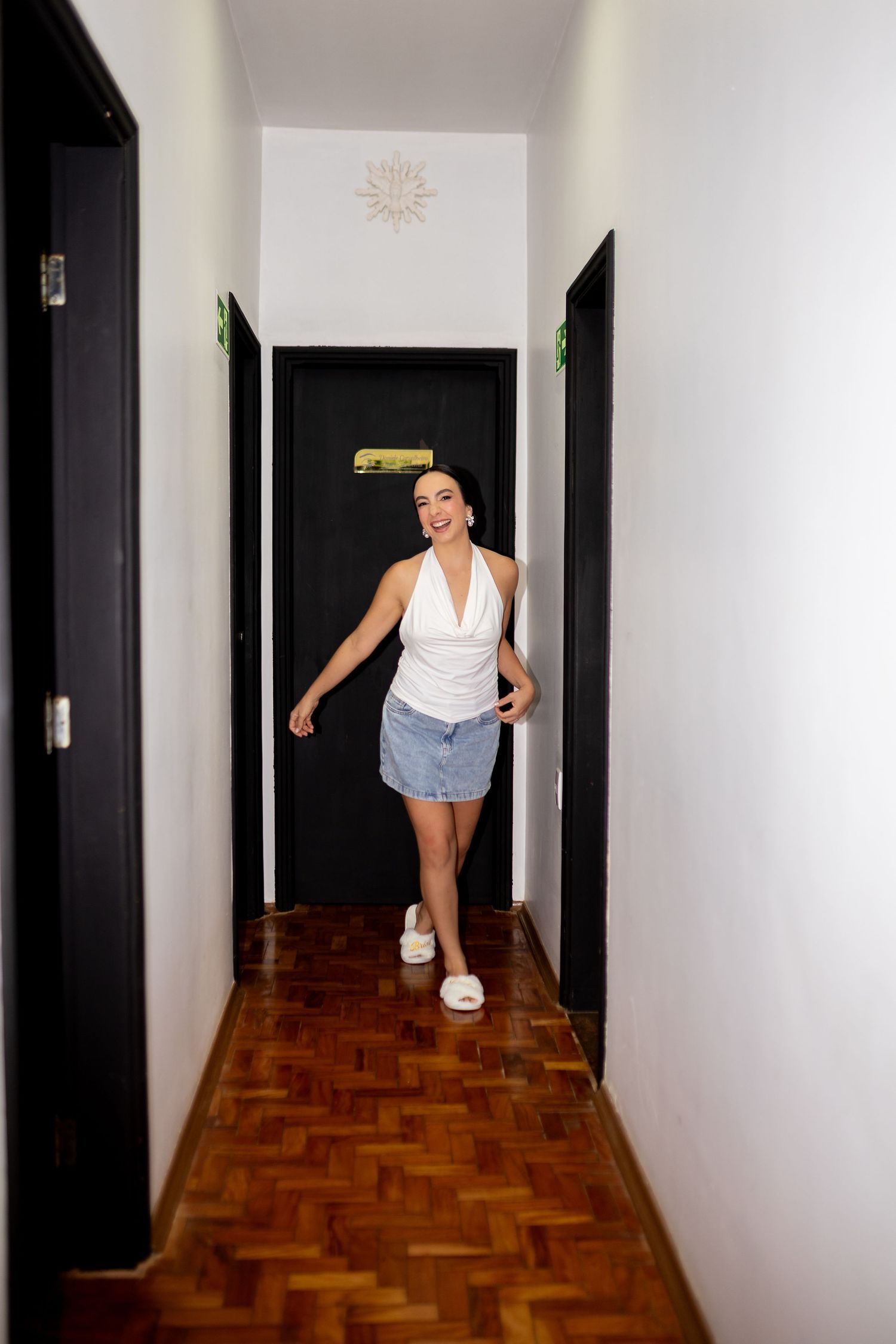 A smiling woman in a white halter top and denim skirt walks through a narrow hallway with dark doors.