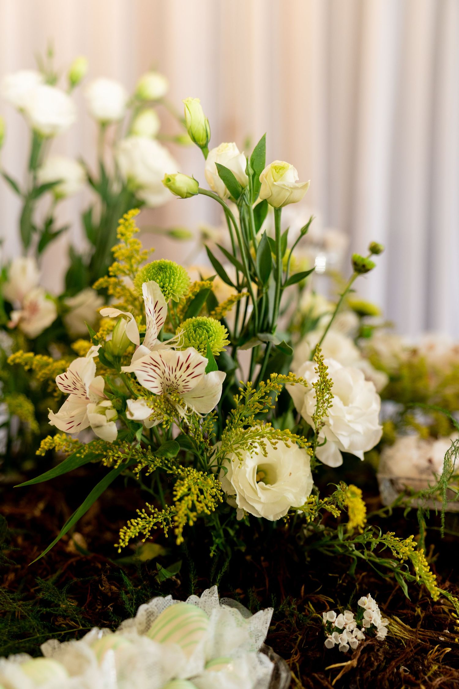 Elegant white and green floral arrangement with ranunculus, alstroemeria, and chrysanthemums on a moss-covered surface.