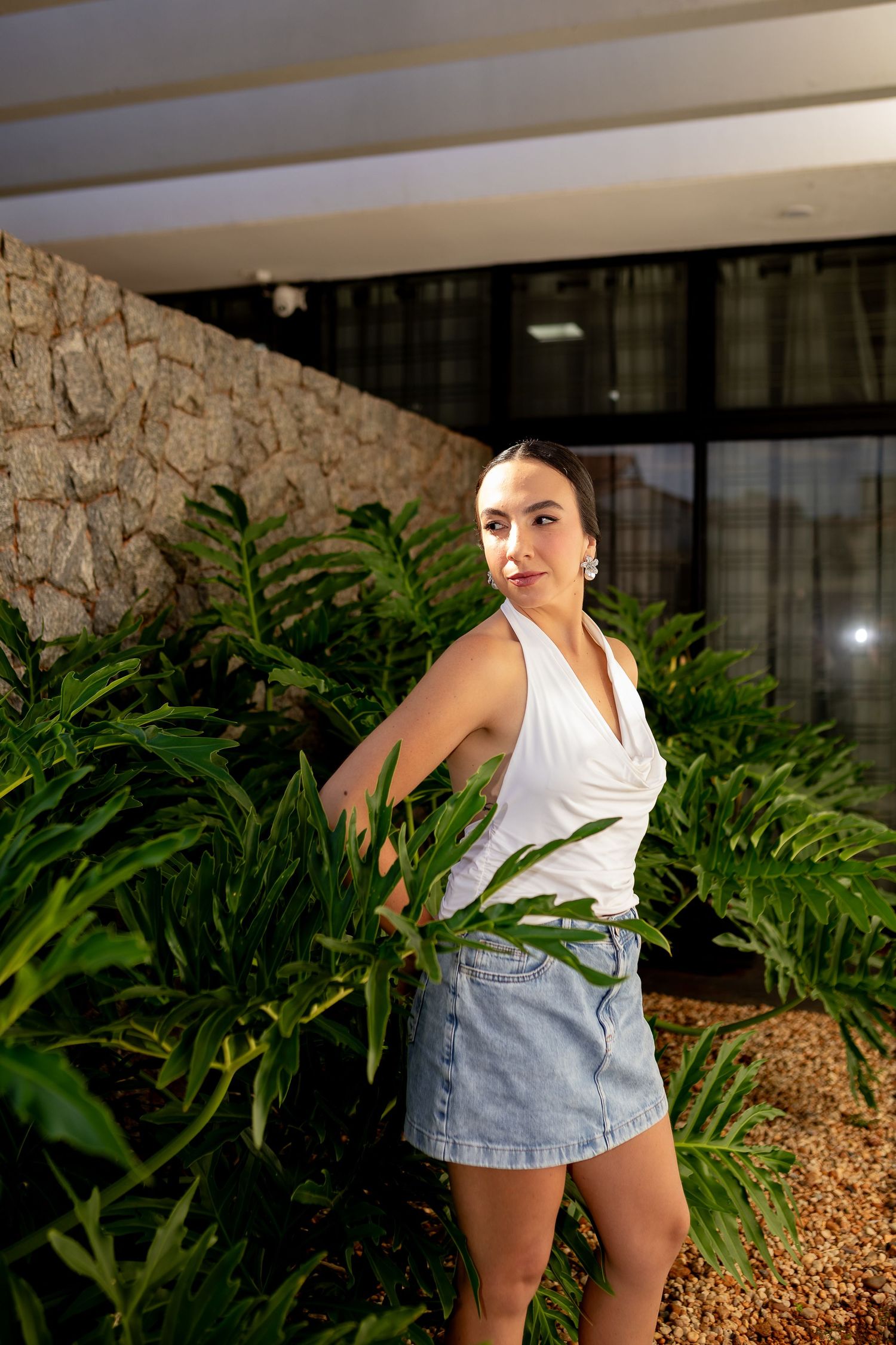 Young woman in white top and denim skirt posing among lush green tropical ferns near stone wall.