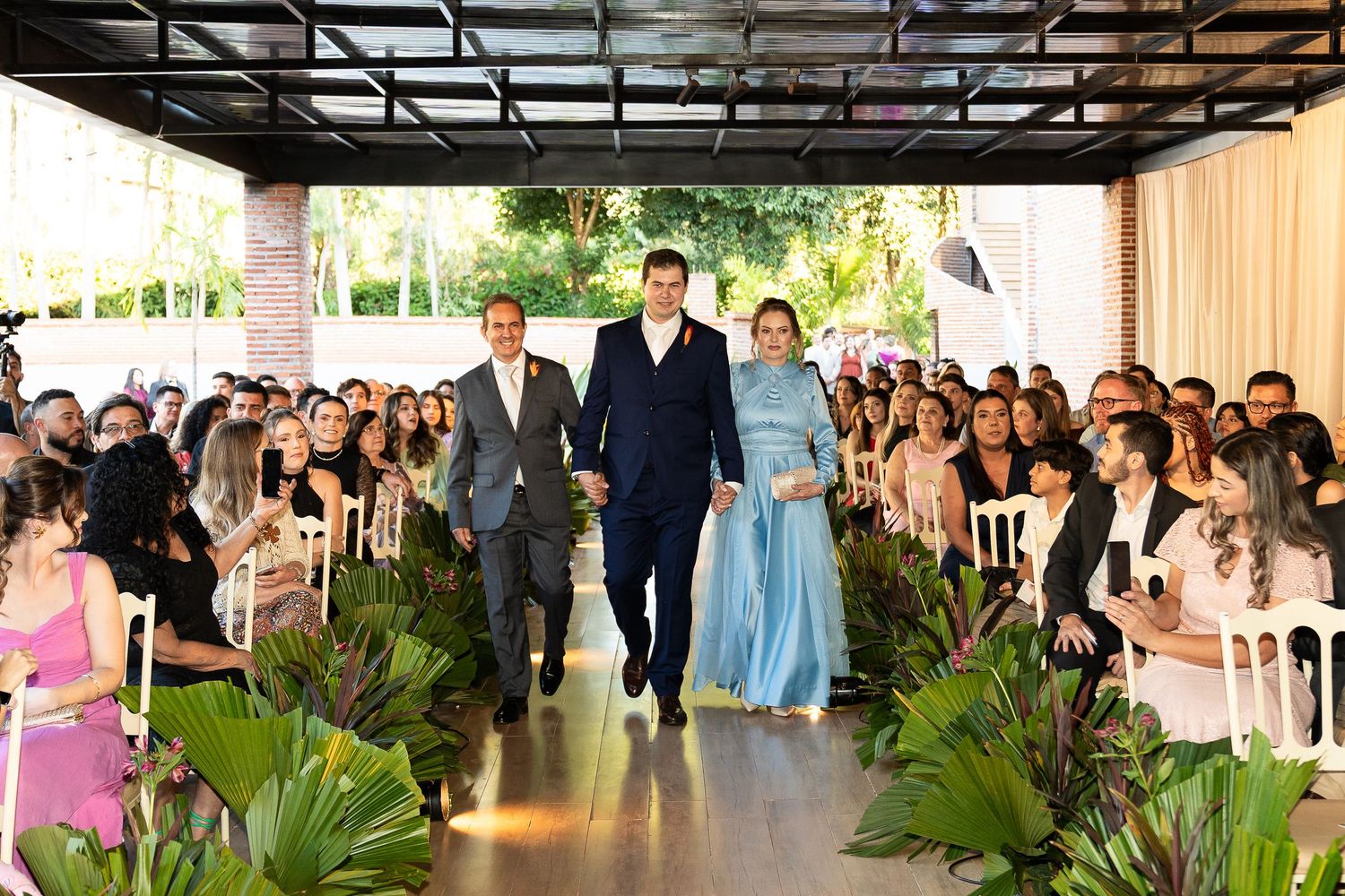 A bride in blue gown walks down a floral aisle with two suited escorts at an elegant outdoor wedding ceremony.