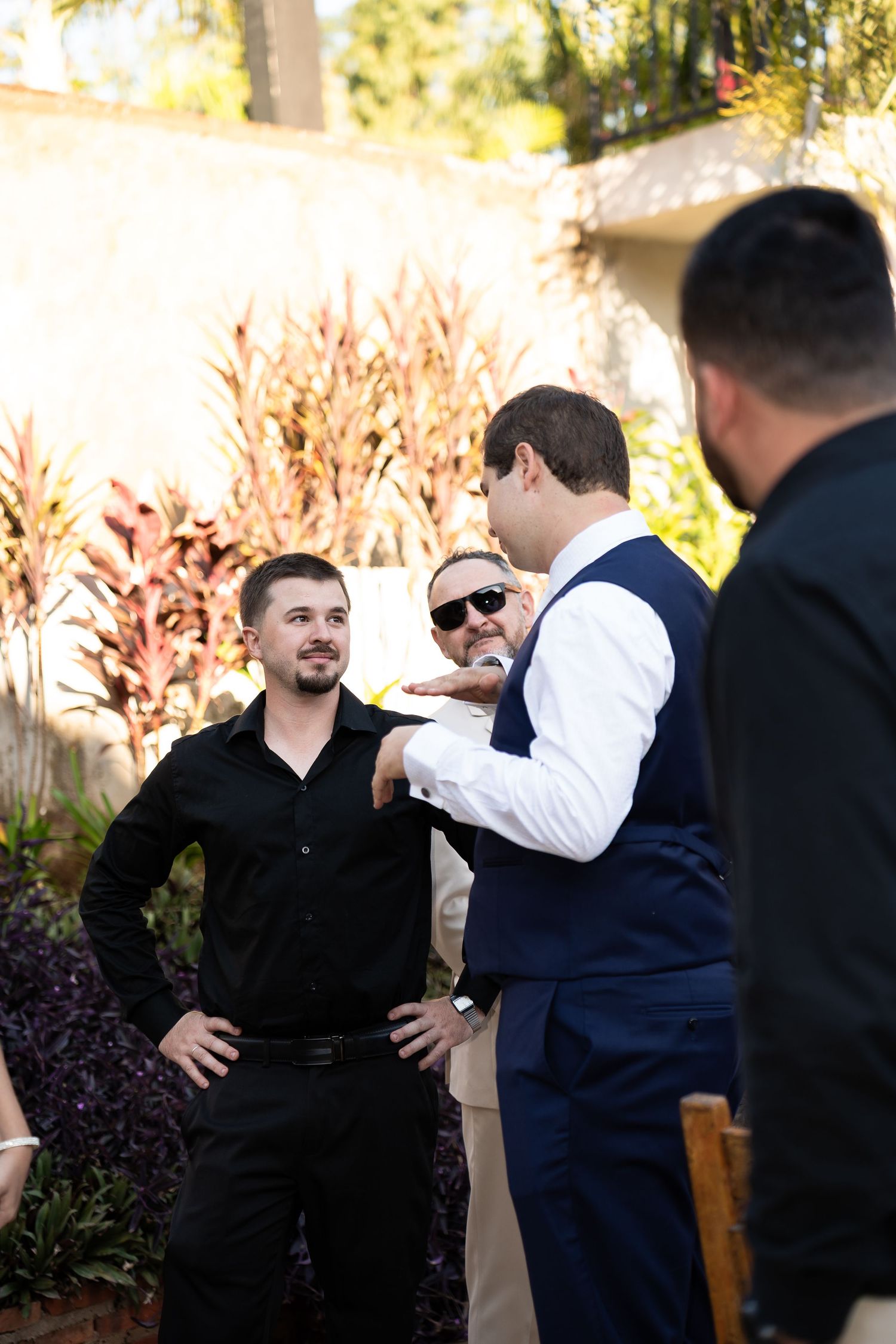 Groomsmen in navy vest and black shirt chat outdoors at a wedding reception surrounded by tropical plants.