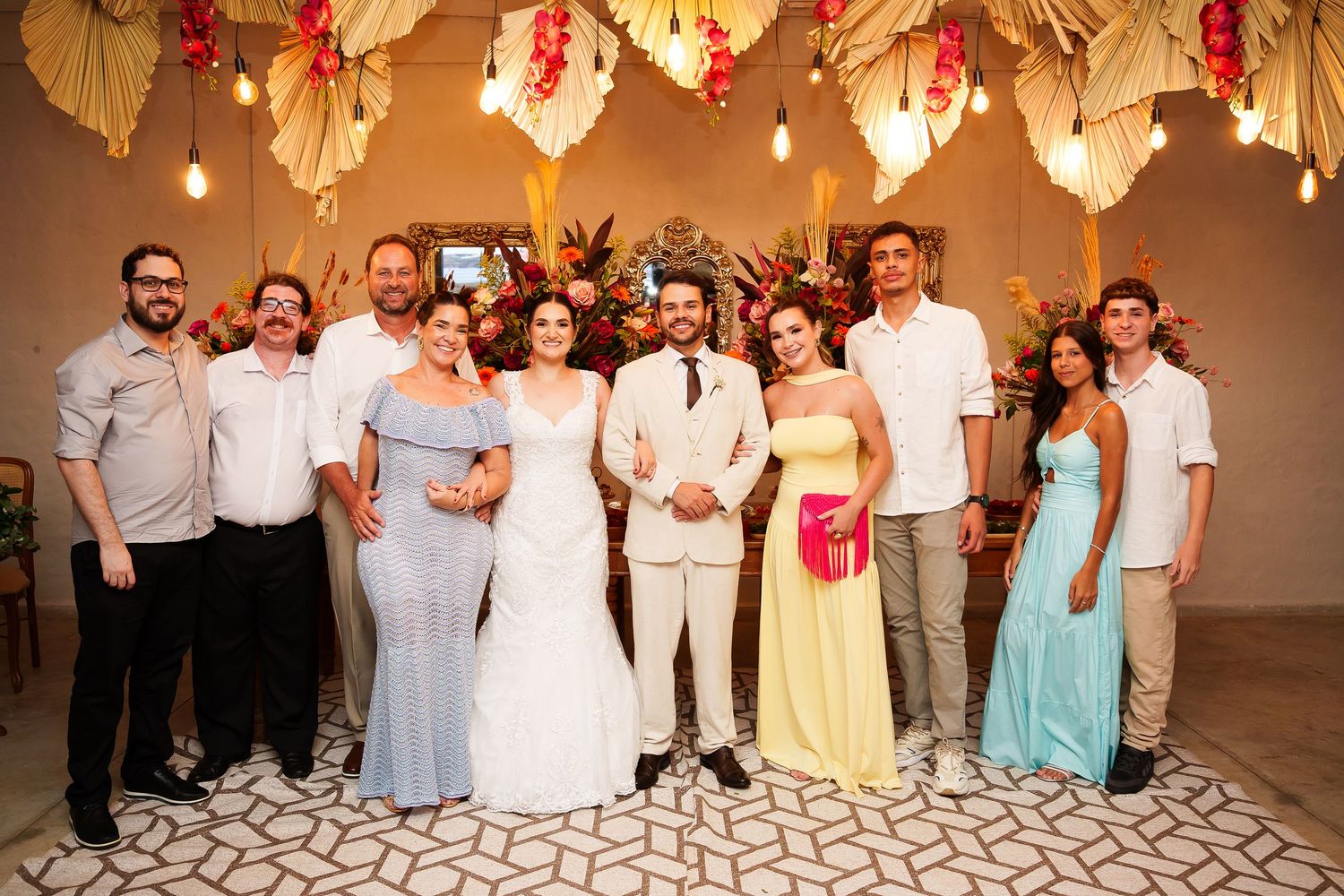Wedding party posing together under floral decorations, bride in white gown and groom in beige suit surrounded by guests.