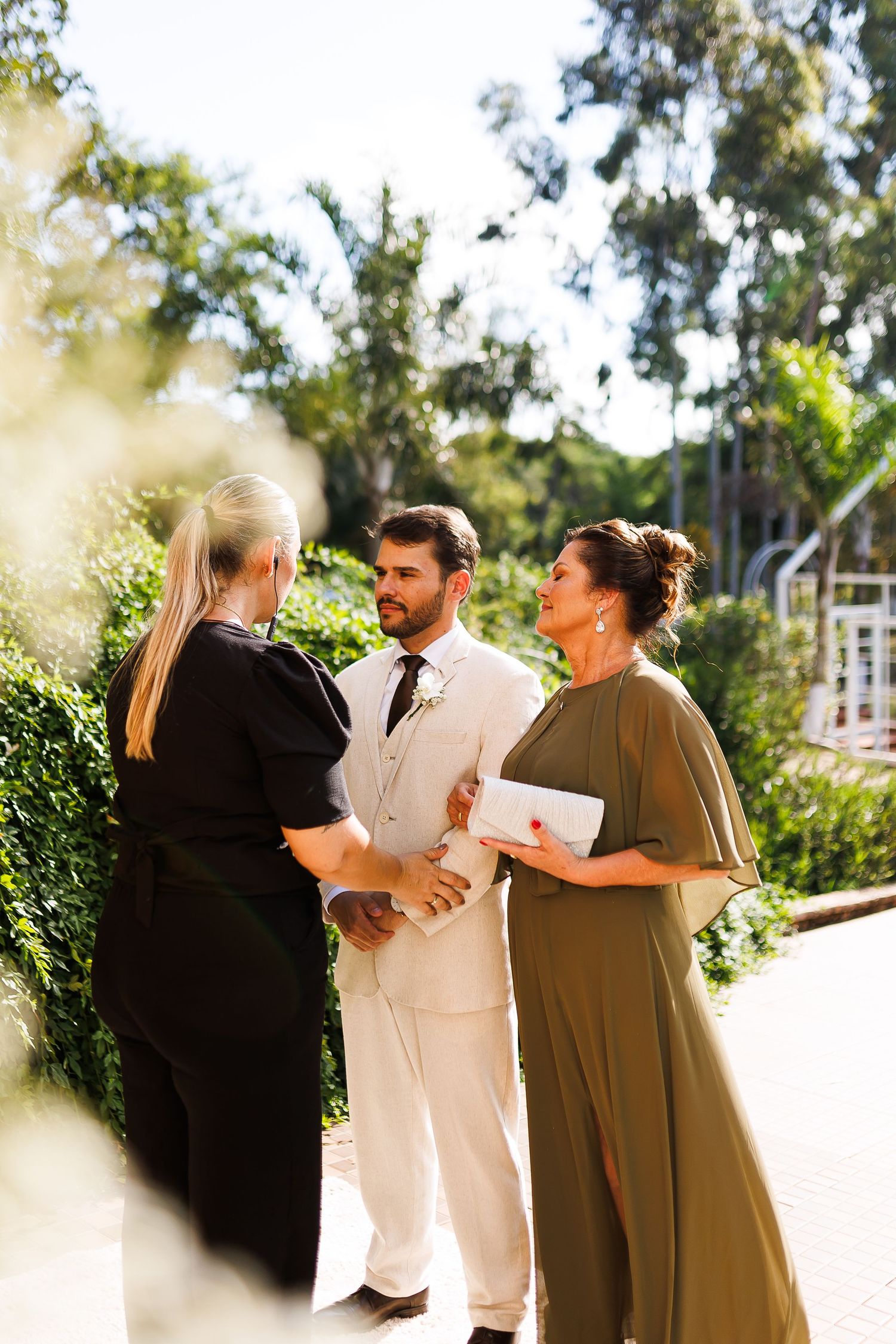 Groom in white suit stands with two women outdoors at a sunny garden wedding ceremony.