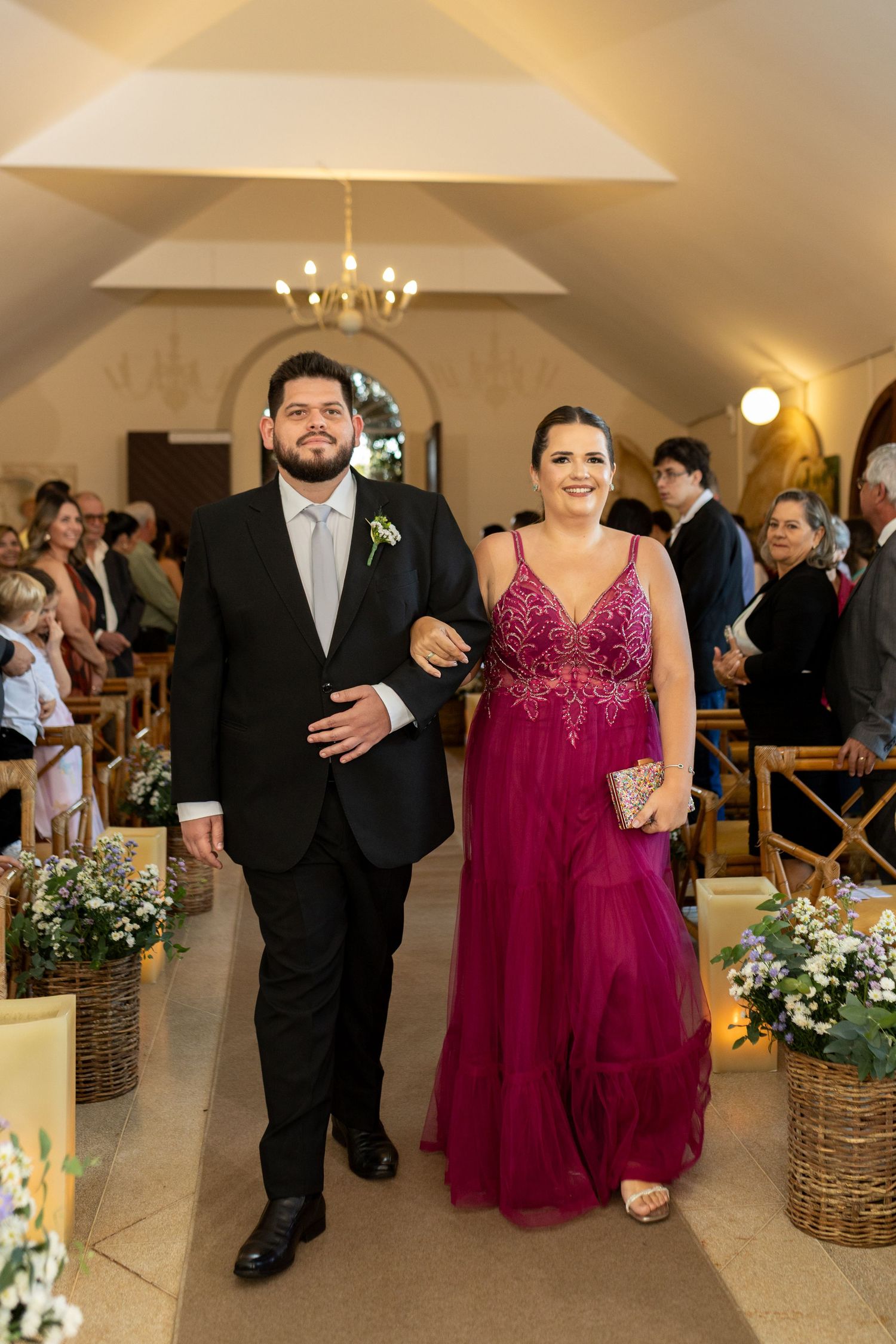 Couple walks down wedding chapel aisle, man in black tuxedo and woman in elegant magenta gown.