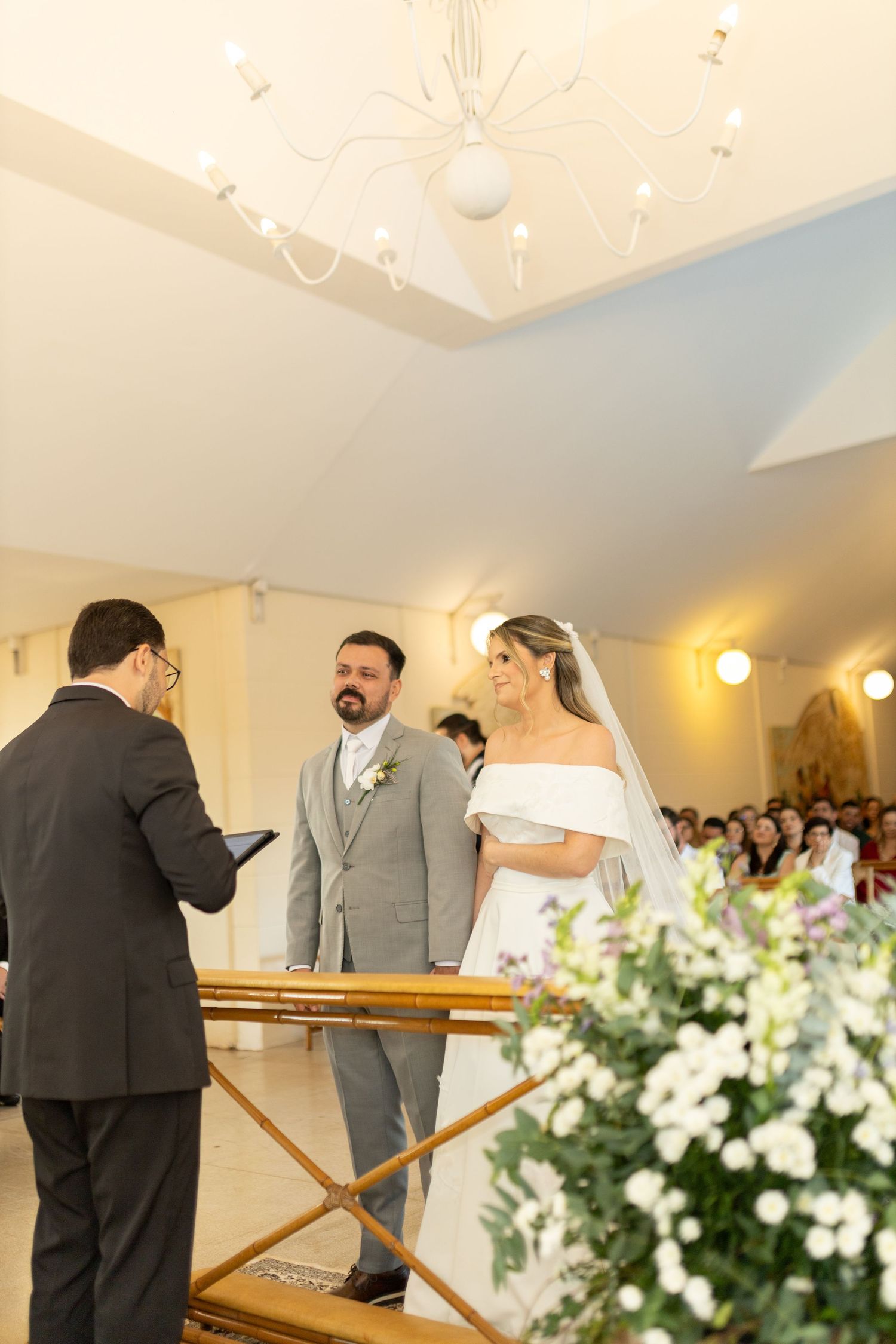 Bride and groom exchange vows at elegant church altar with floral arrangements and chandelier during wedding ceremony.