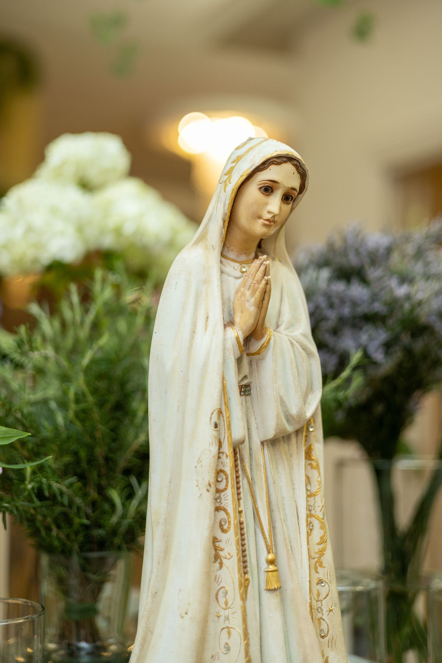 White ceramic Virgin Mary statue with hands in prayer, surrounded by fresh flowers and soft bokeh lighting.