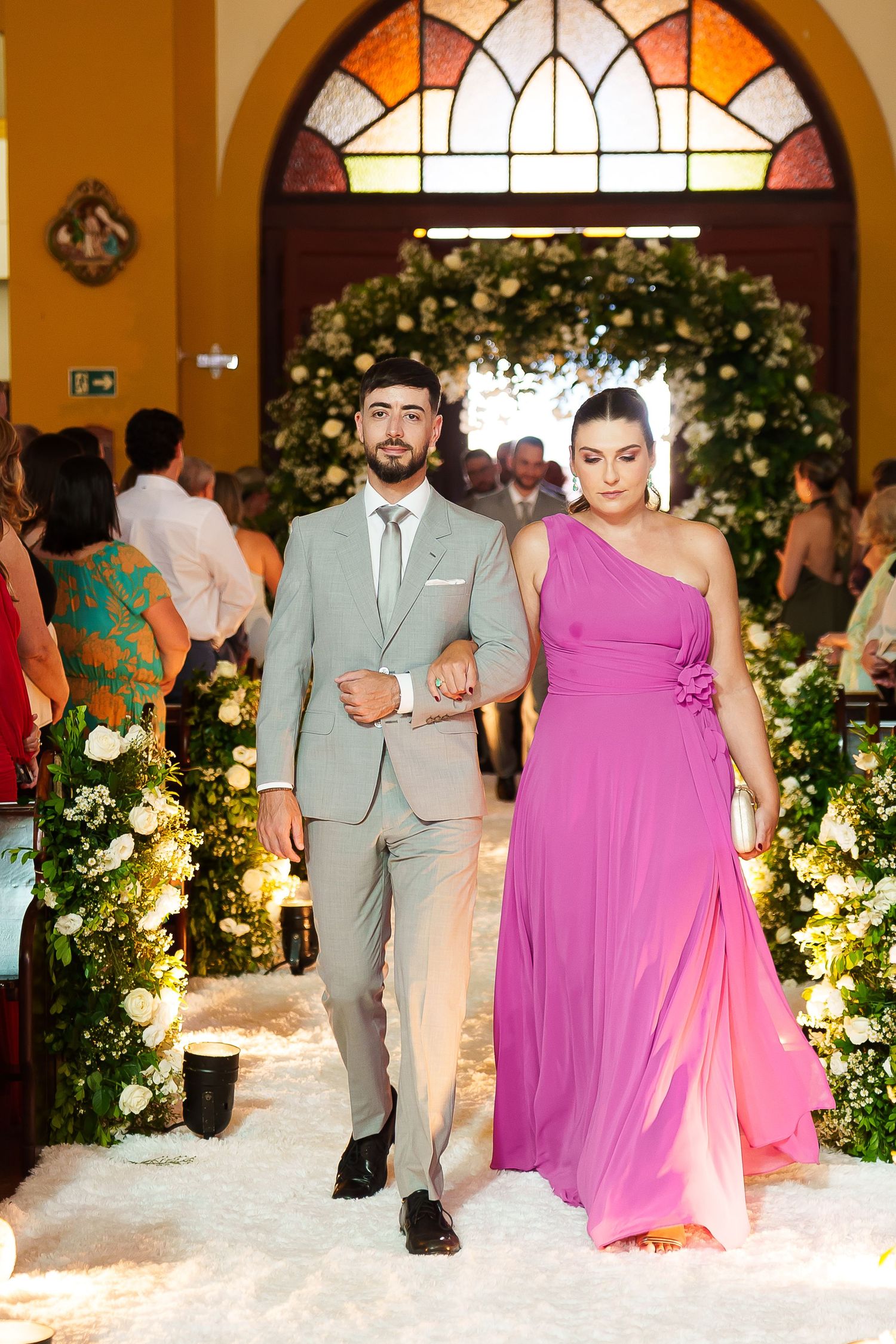 A couple walks down the aisle at a wedding, man in gray suit and woman in a purple one-shoulder gown.