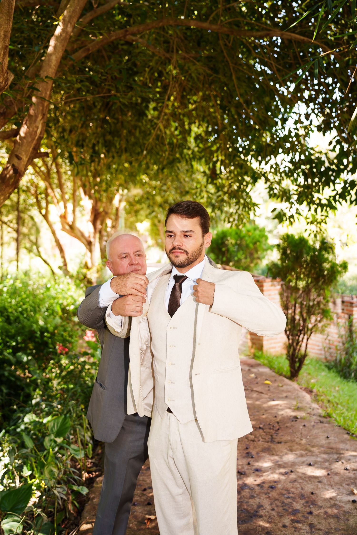 Two men in suits outdoors, older man adjusting younger groom's jacket collar in a garden setting.