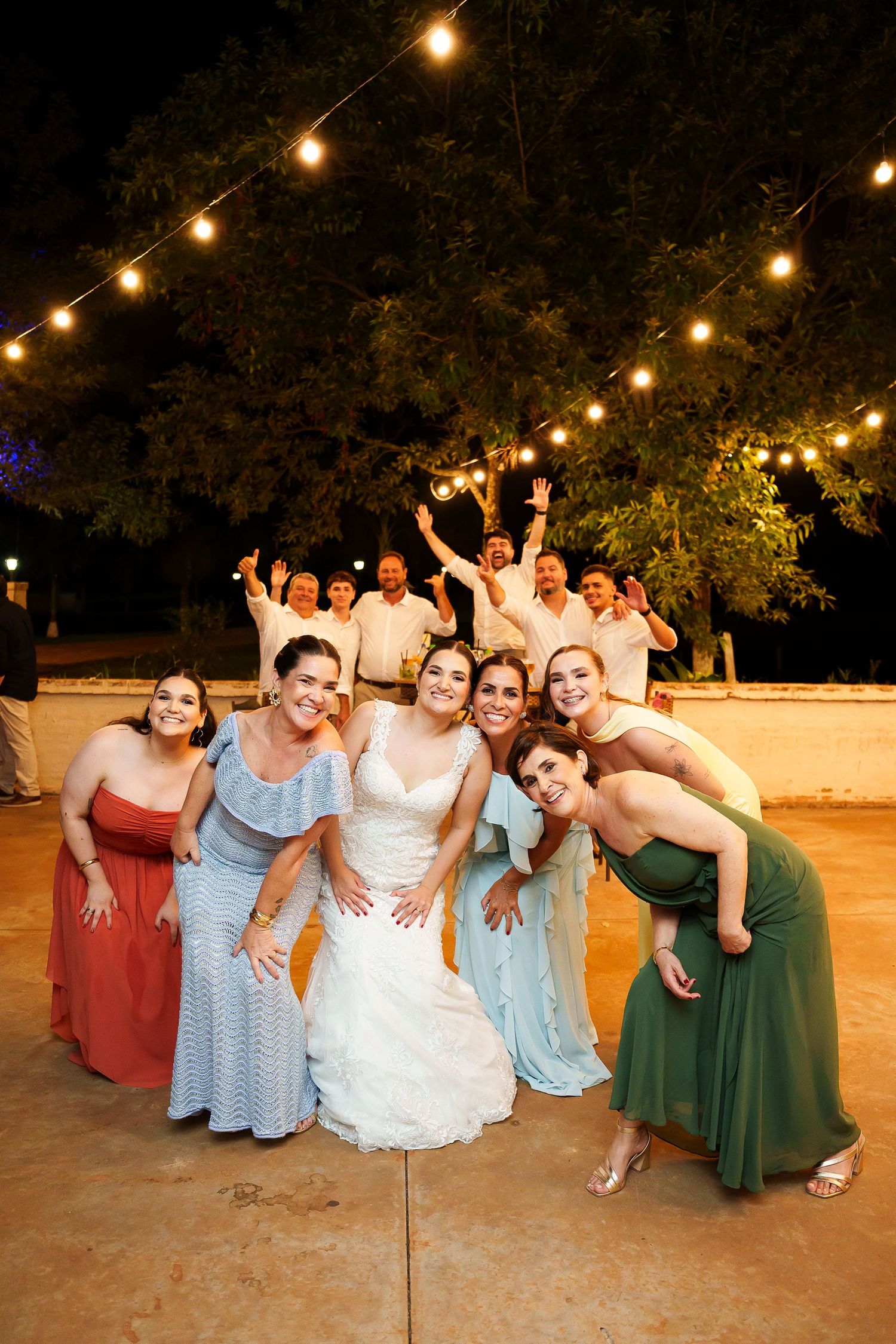 Bride poses with bridesmaids in colorful gowns at outdoor wedding reception under string lights at night.