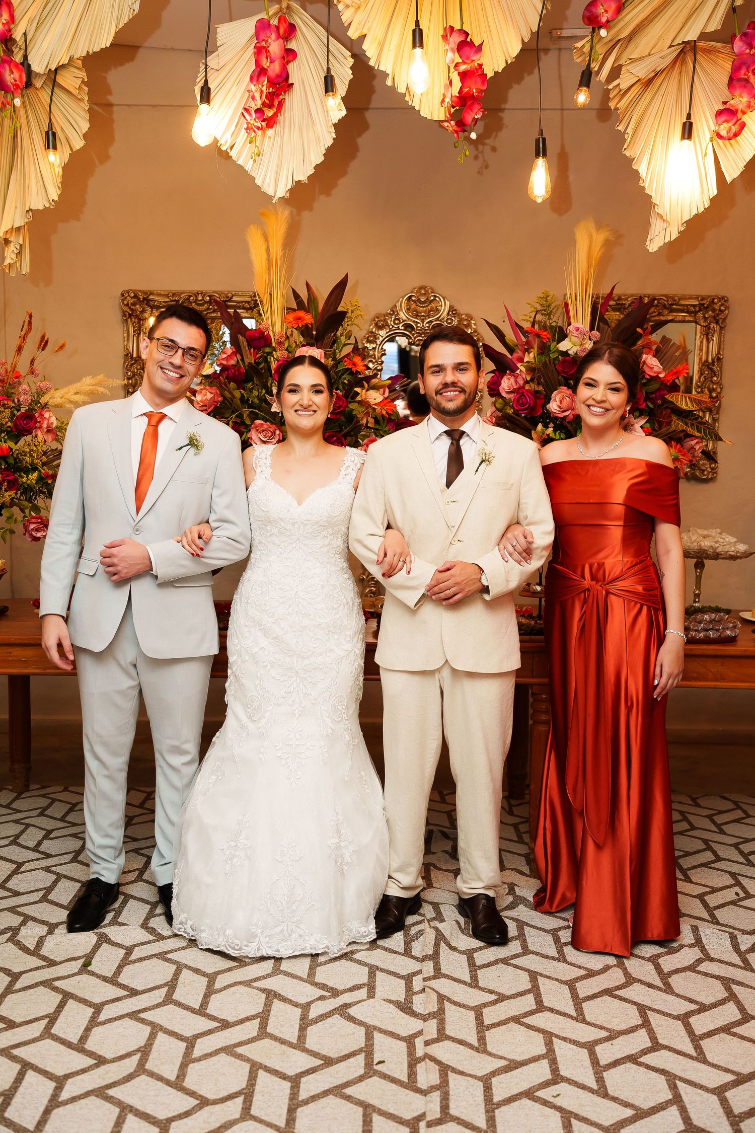 Wedding party of four posing elegantly, bride in white gown, groom in cream suit, guests in orange and gray formal attire.