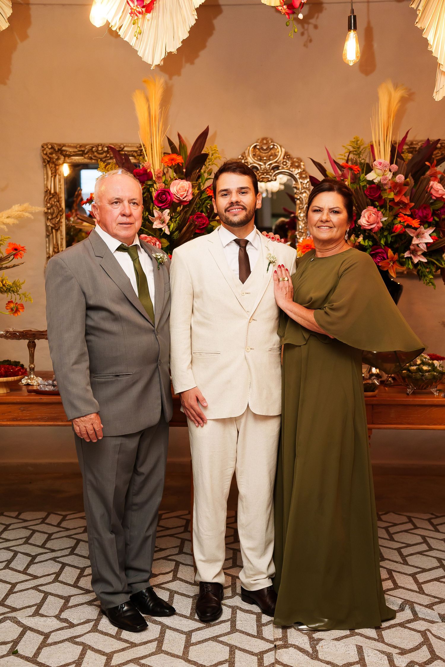 A groom in a white suit poses with his parents at a wedding reception, surrounded by colorful floral arrangements.