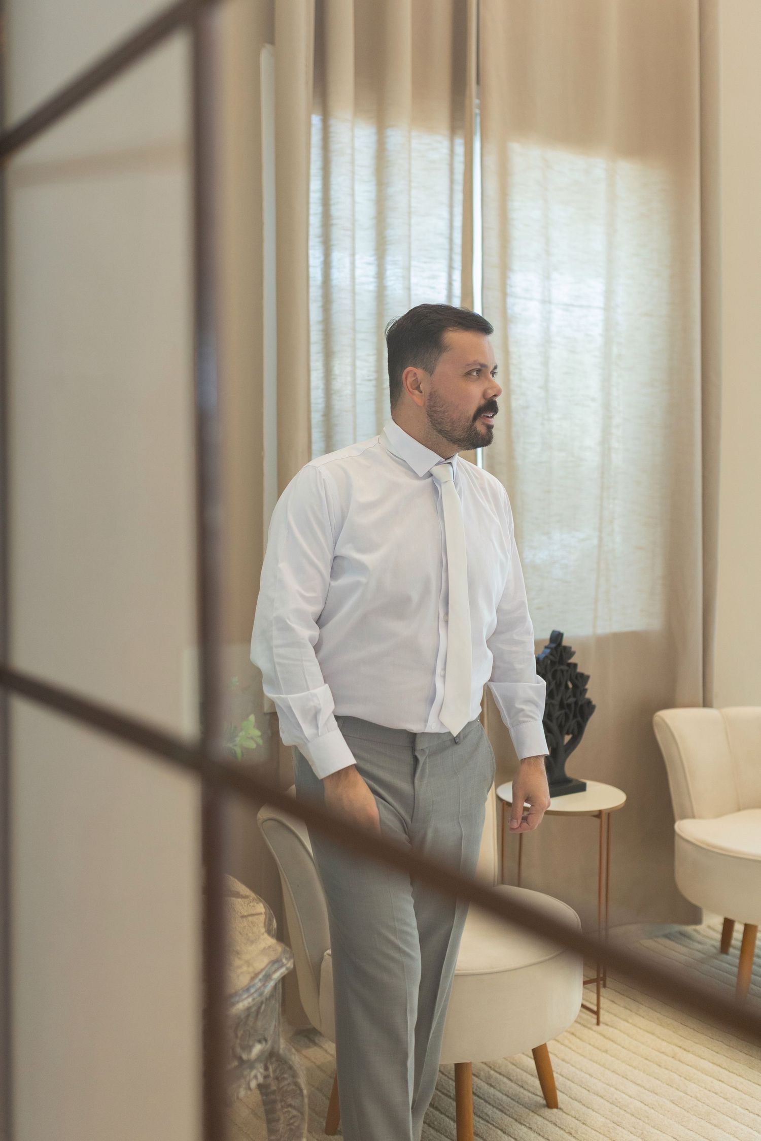 A well-dressed man in a white shirt and gray pants stands thoughtfully by a bright window in an elegant room.