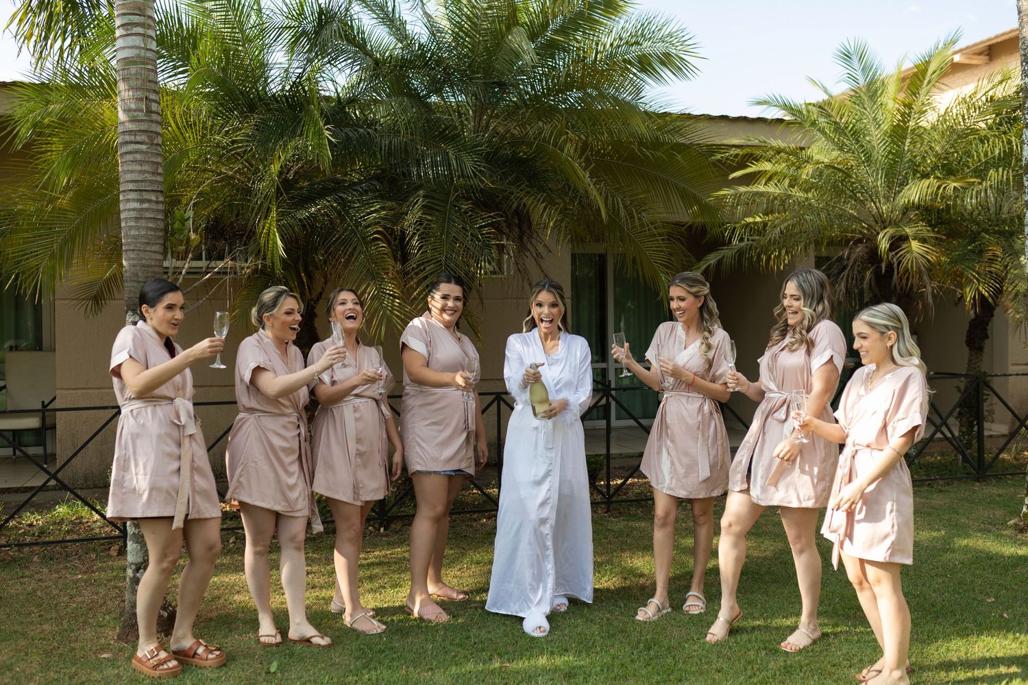 Bride in white robe celebrates with bridesmaids in matching pink robes outdoors near tropical palm trees.