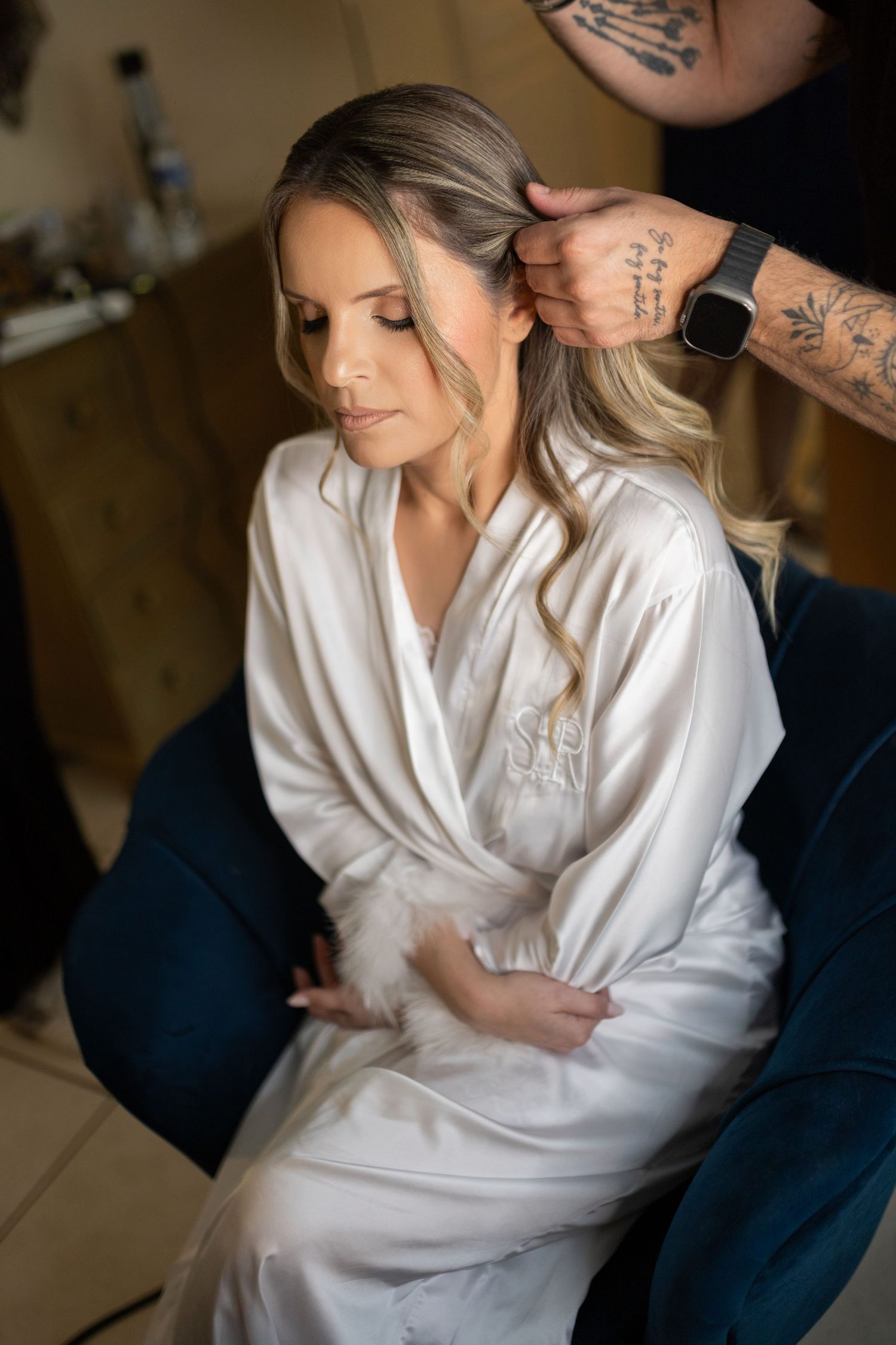 Bride in white satin robe gets hair styled by tattooed hairstylist during wedding morning preparations.