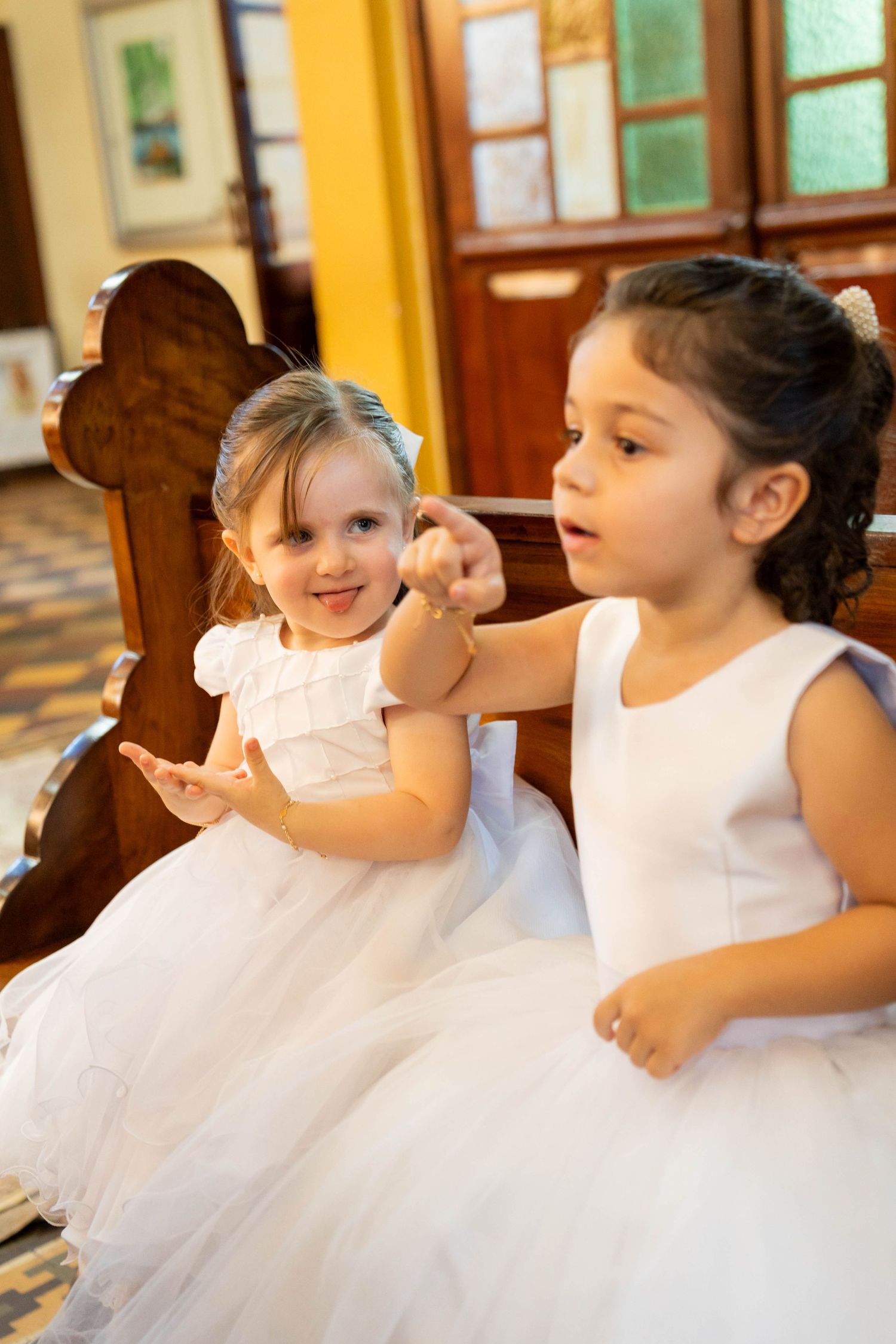 Two young girls in white dresses sitting together in a church pew, one pointing and smiling.