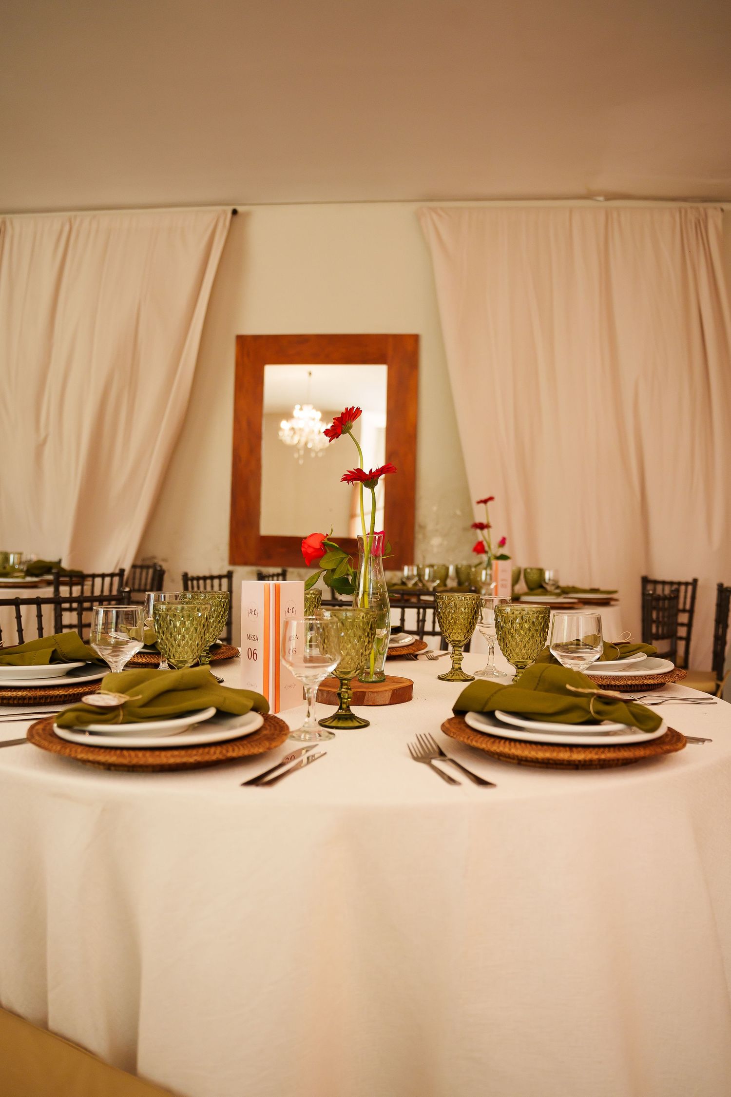 Elegant wedding reception table with green goblets, wicker chargers, white linens, and red floral centerpiece in a draped tent.