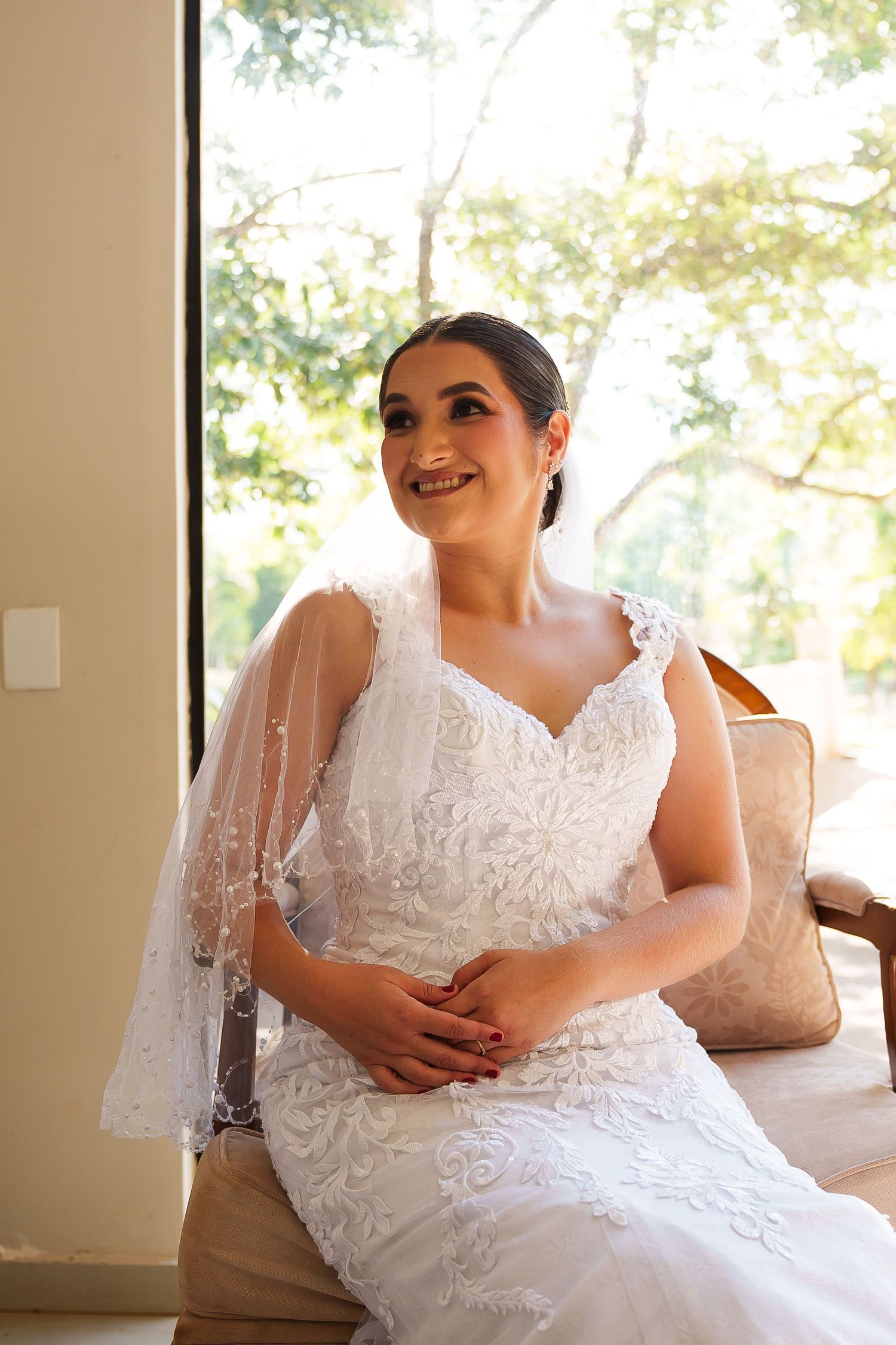Smiling bride in white lace wedding dress and veil sits gracefully near a sunlit window with trees in background.