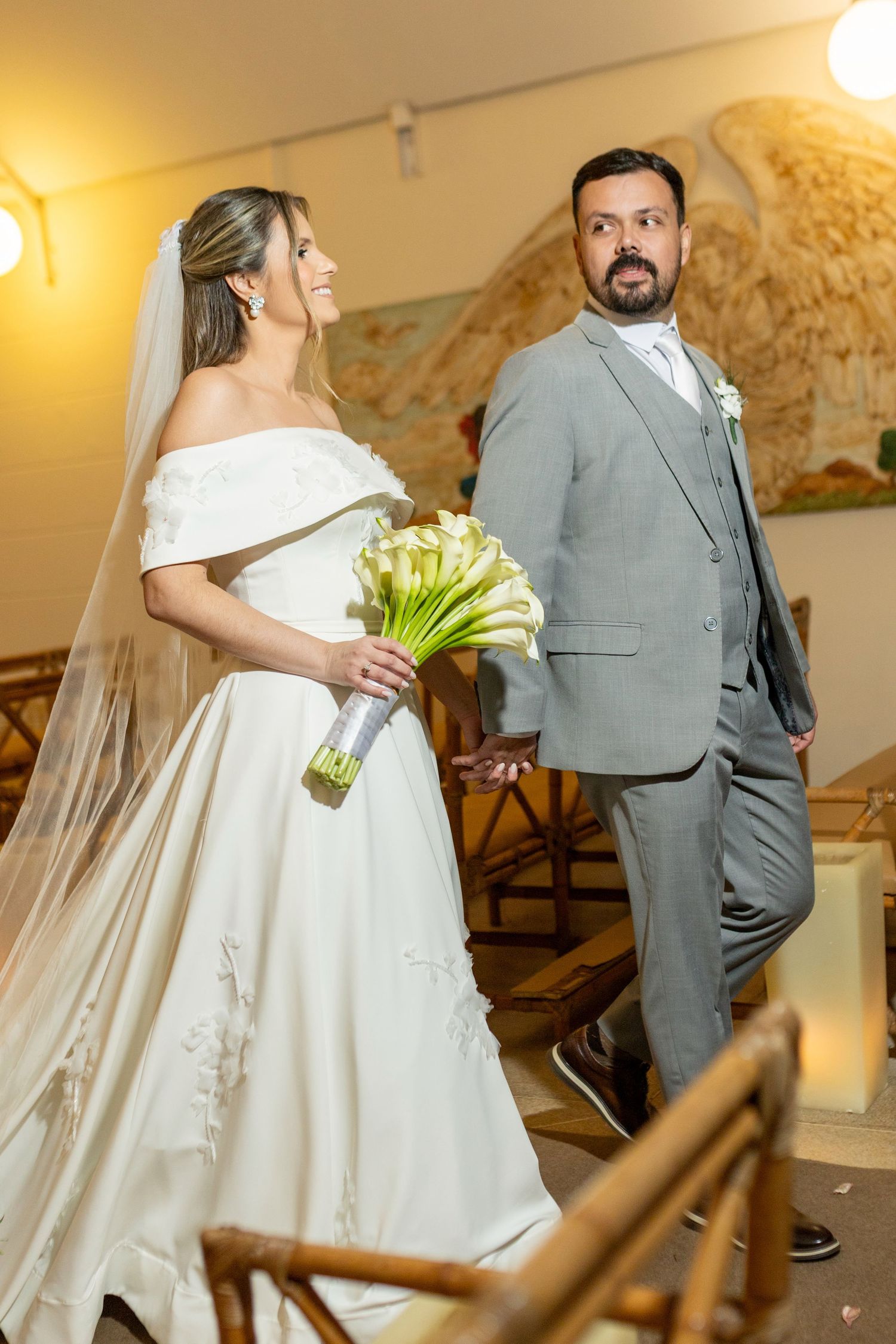 Bride in white off-shoulder gown holding calla lily bouquet stands with groom in gray suit at church wedding ceremony.