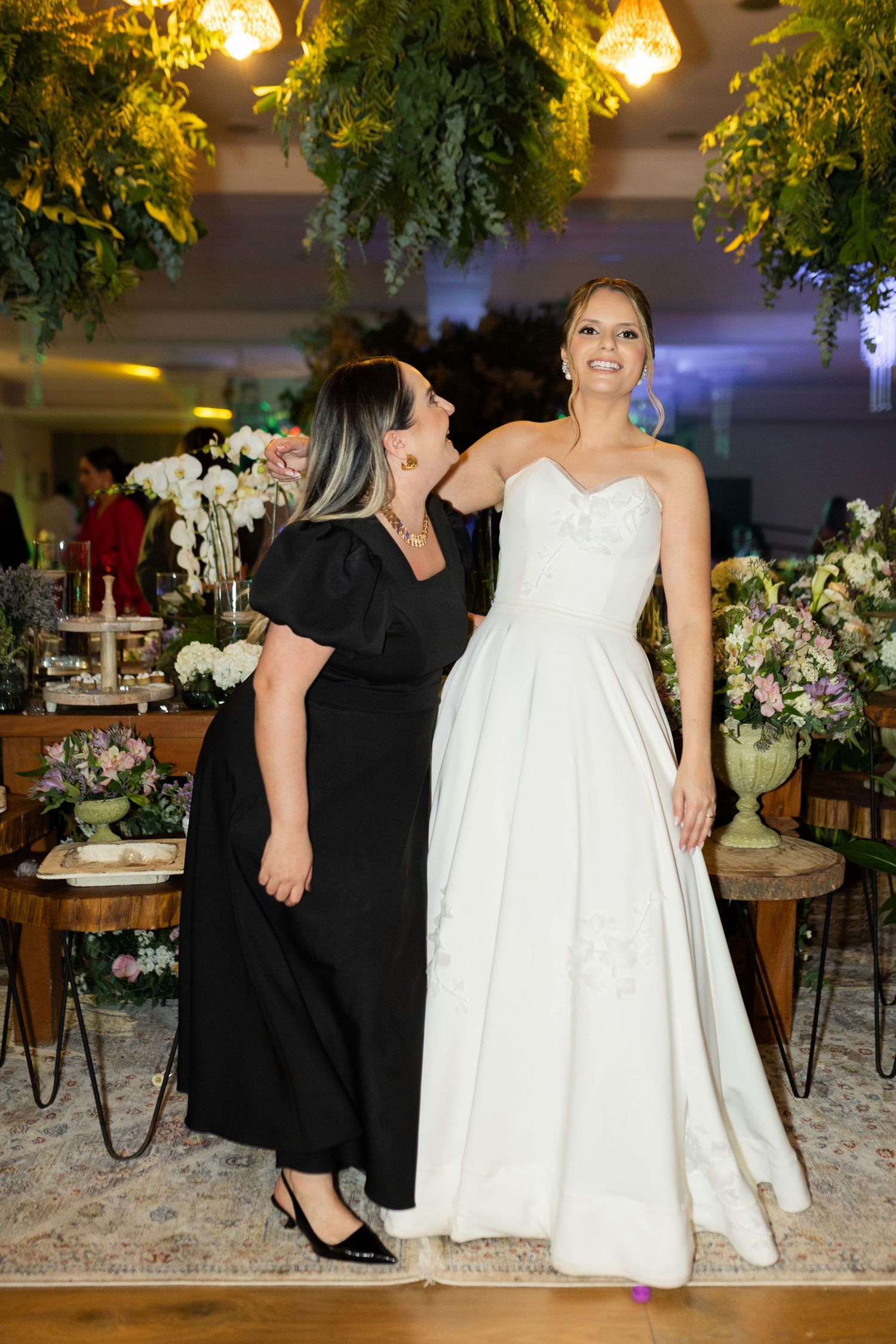 Bride in white strapless gown smiles beside woman in black dress at elegant floral wedding reception.