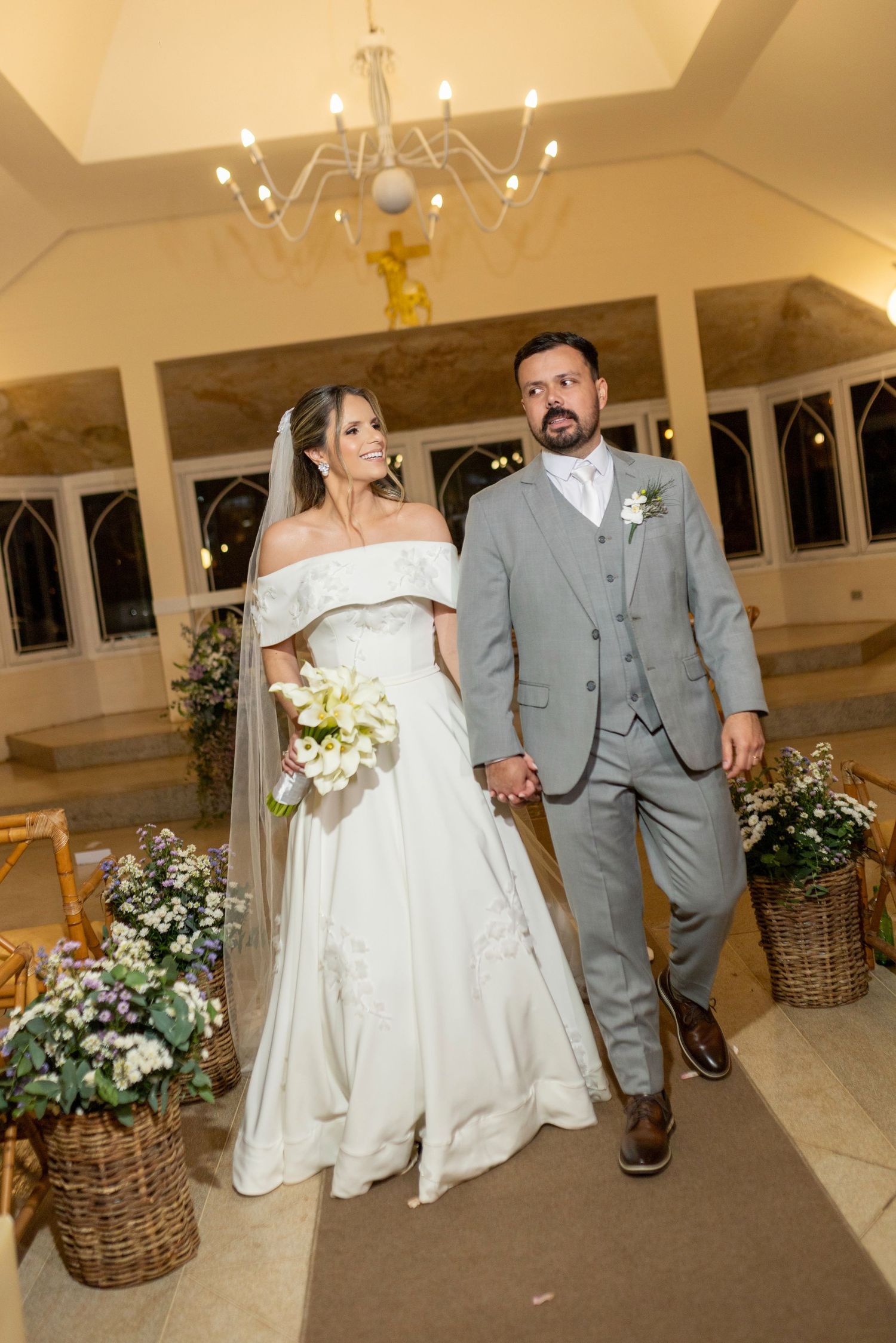 Bride and groom walk down church aisle, she in white off-shoulder gown with veil, he in gray suit.