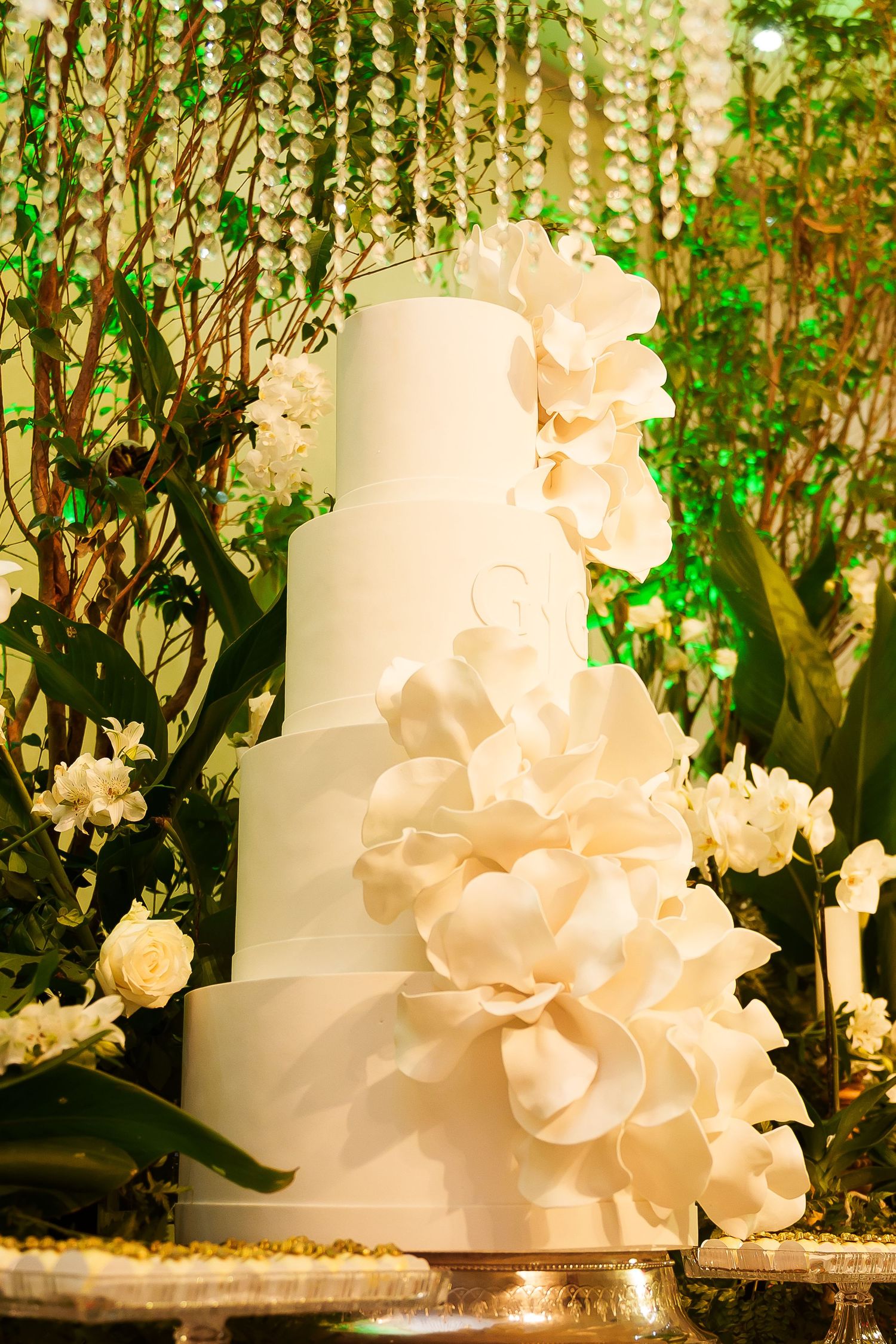 Elegant five-tier white wedding cake adorned with large sugar flowers, displayed against a lush green floral backdrop.