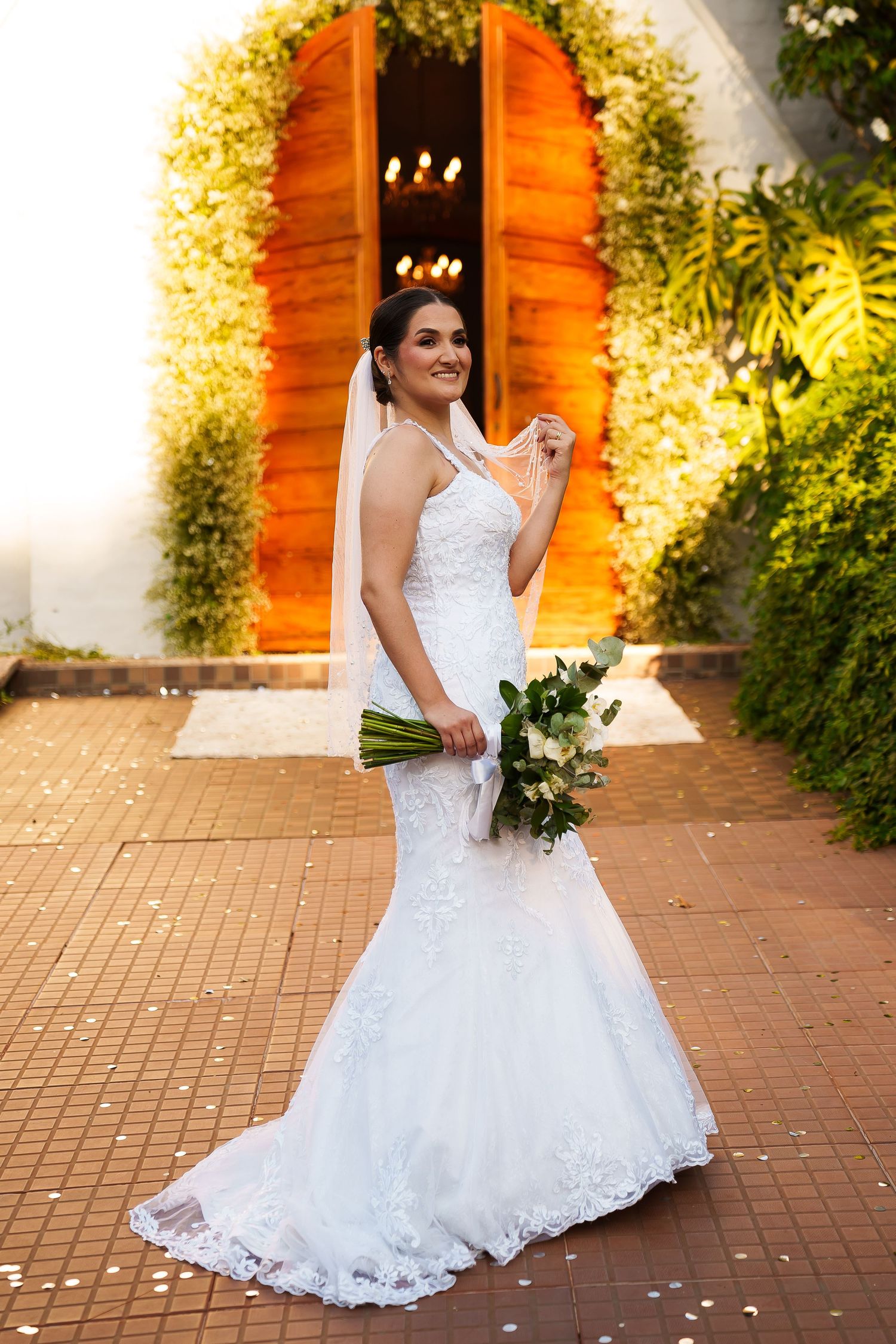 Smiling bride in white lace mermaid gown holding bouquet, standing before ornate wooden door with floral greenery.