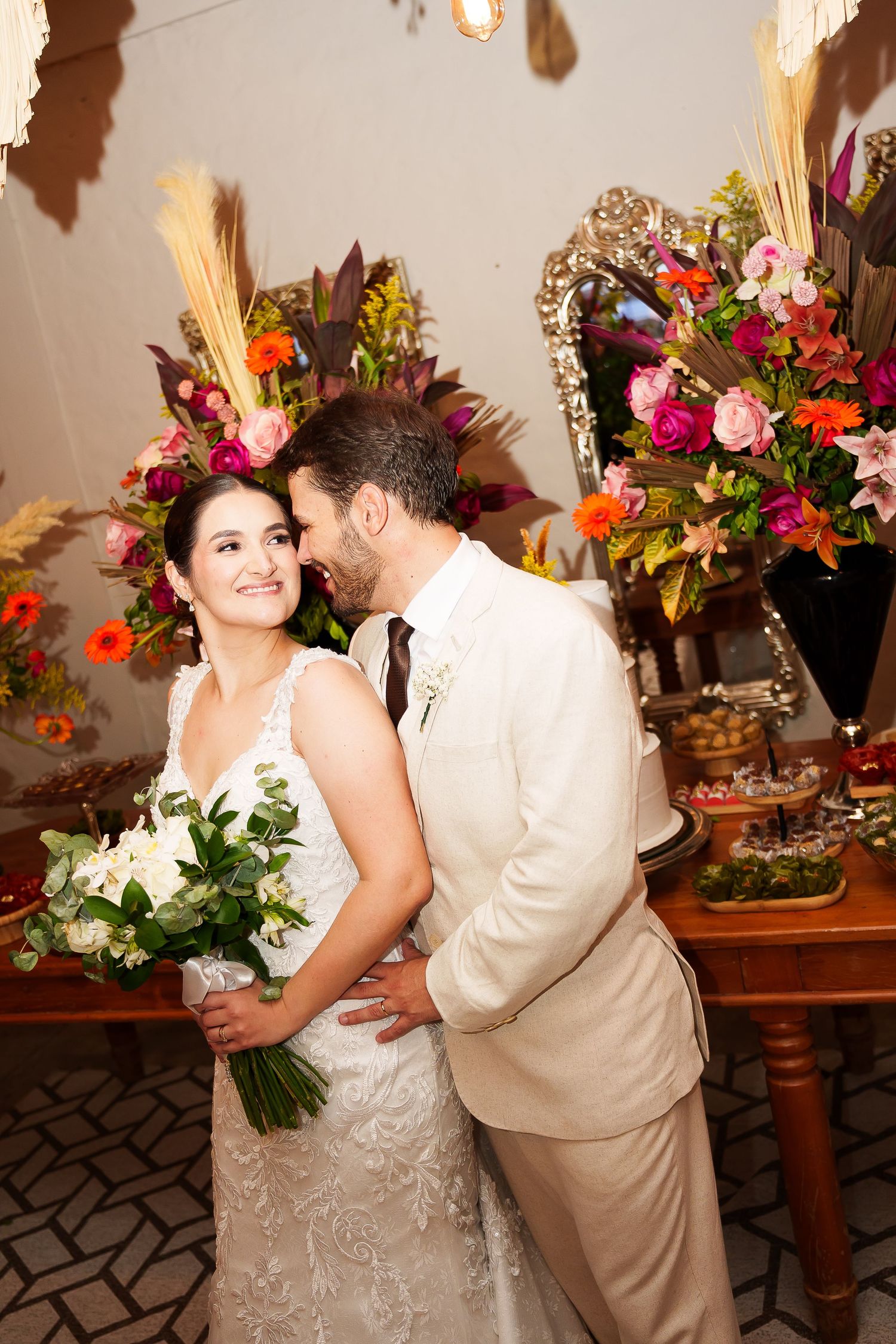 Bride in white lace gown holding bouquet while groom in cream suit kisses her cheek at floral wedding reception.