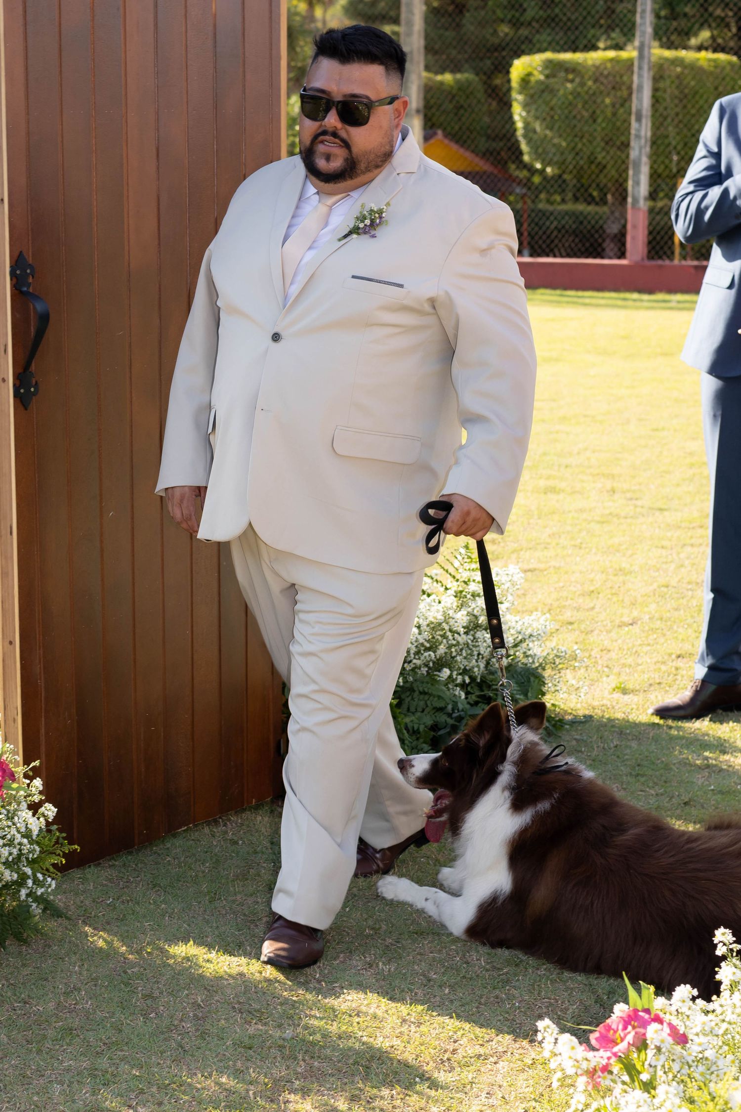 Man in cream suit with sunglasses holds leash of brown and white Border Collie at outdoor wedding.