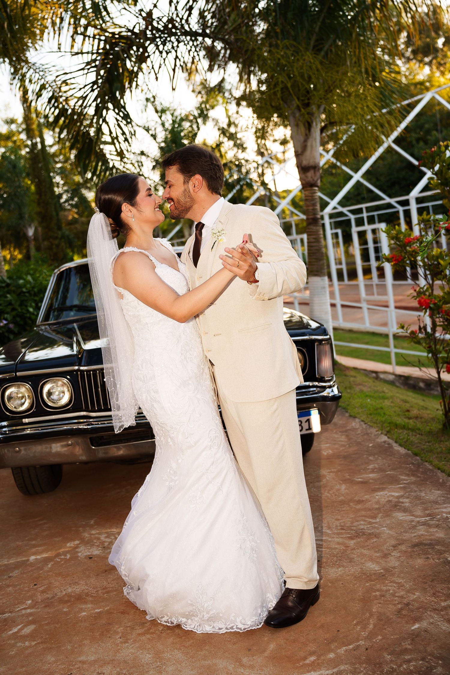 Bride and groom share first dance outdoors near vintage black car, surrounded by palm trees at golden hour.