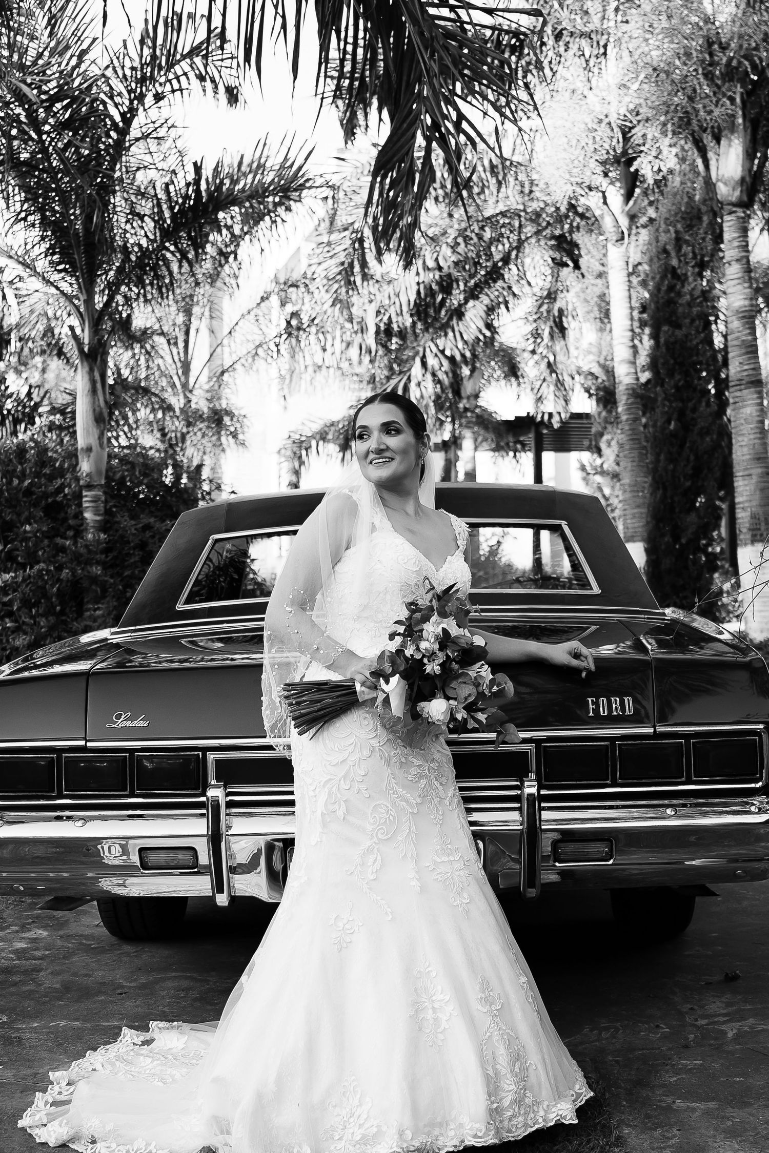 Bride in lace wedding dress and veil holding bouquet poses beside vintage Ford car with palm trees in black and white photo.