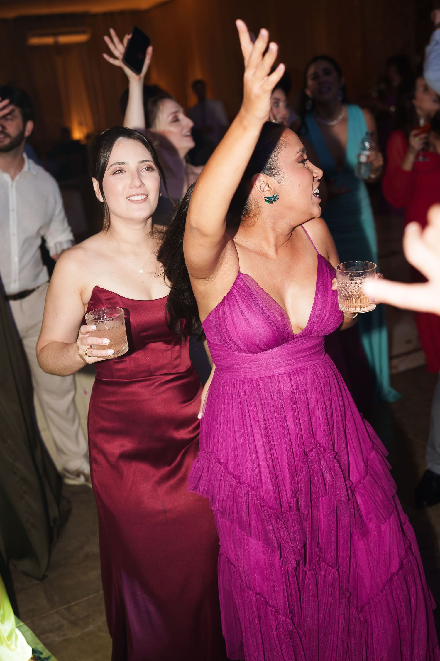Two women in elegant gowns, one burgundy and one magenta, dancing joyfully at a formal event.