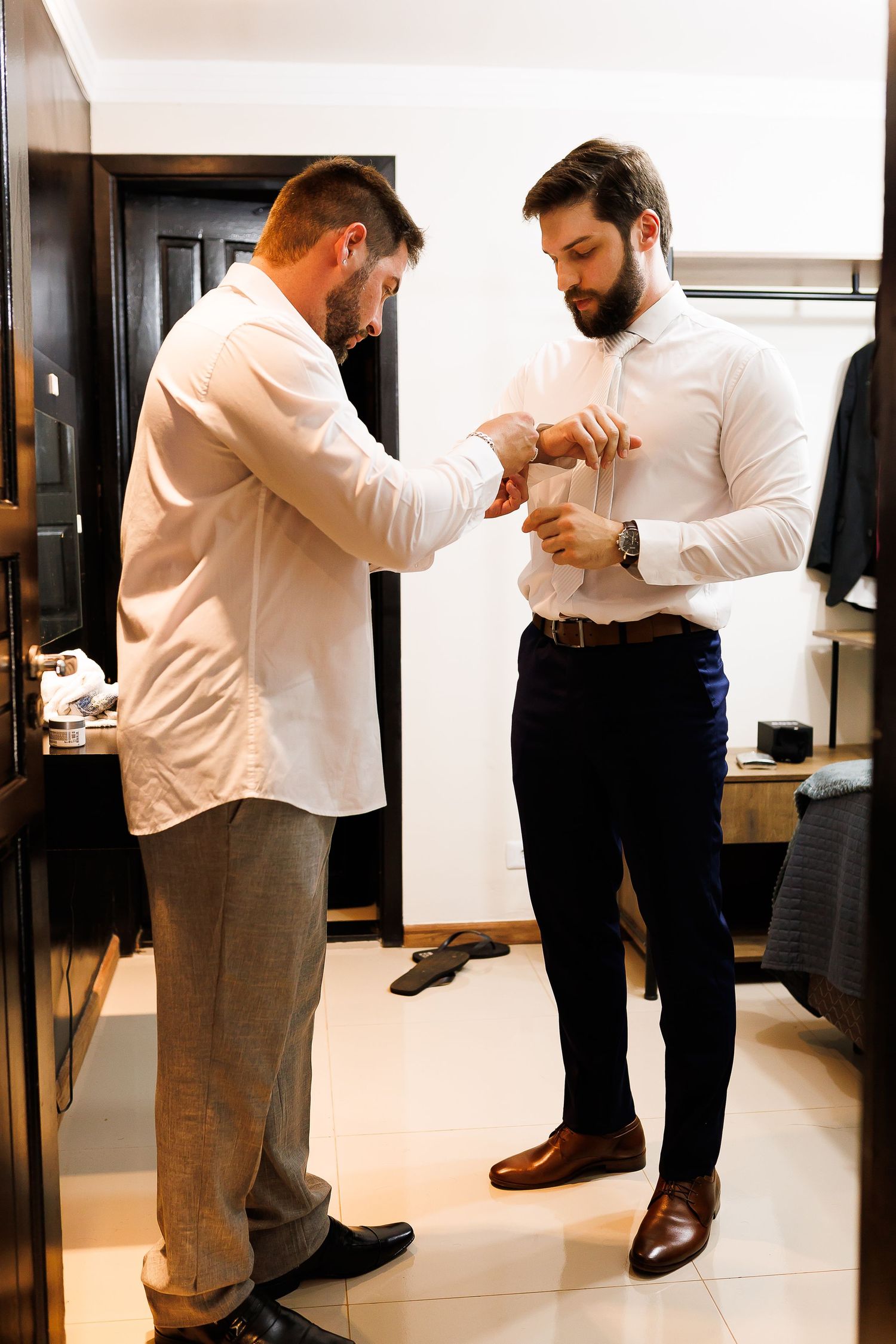 Two well-dressed bearded men in white shirts helping each other with cufflinks in a stylish dressing room.