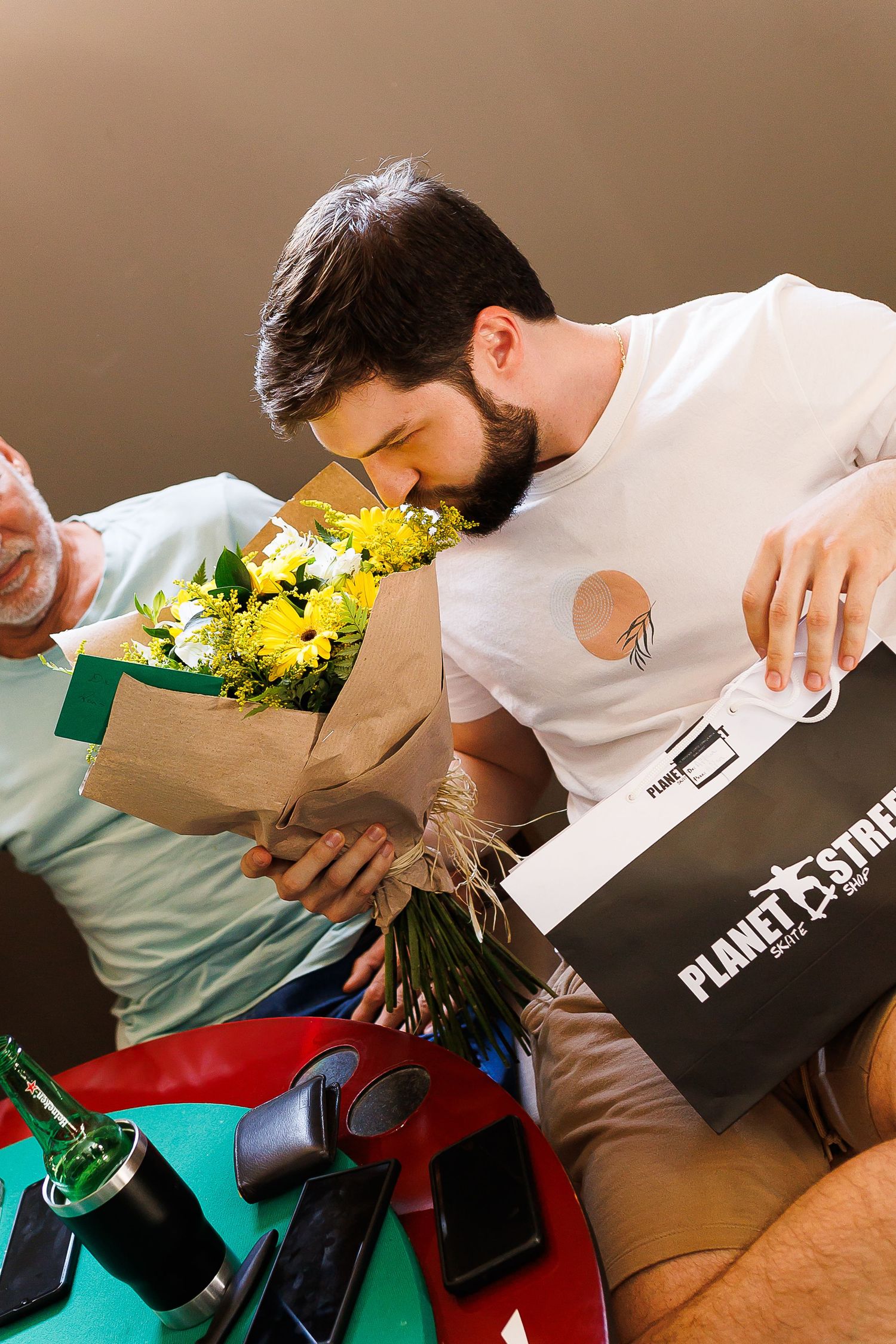 Bearded man smelling yellow flower bouquet while holding a Planet Fitness shopping bag indoors.