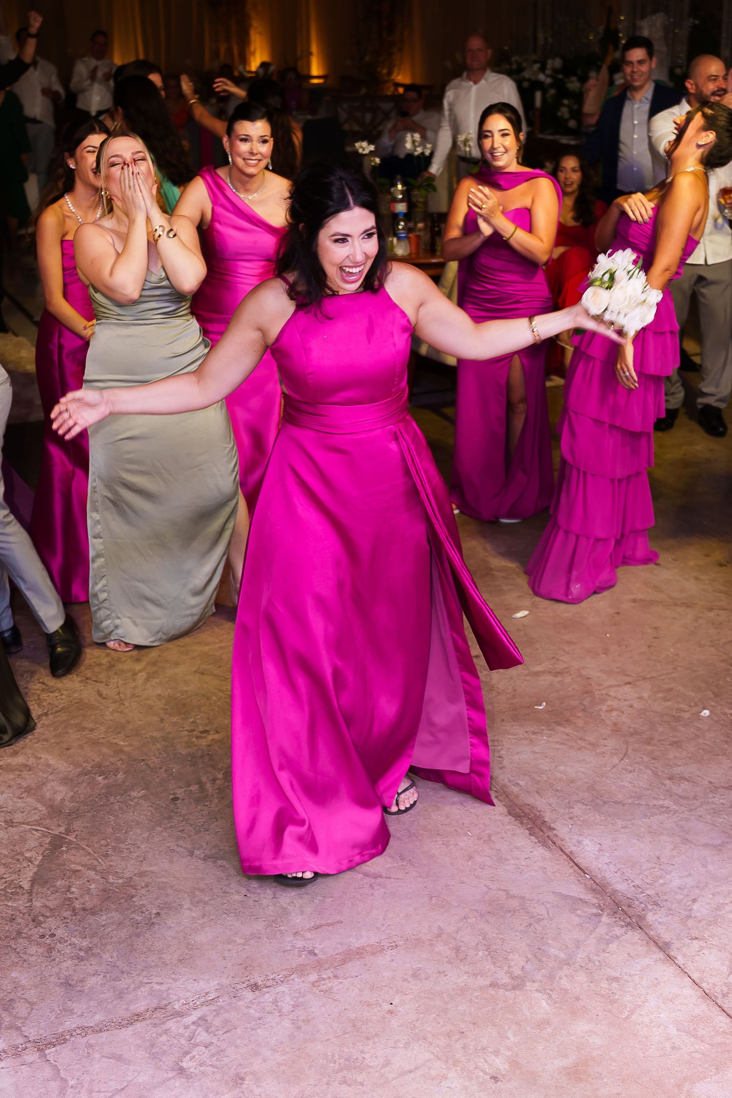 A woman in a fuchsia gown catches the bridal bouquet at a wedding reception surrounded by bridesmaids.