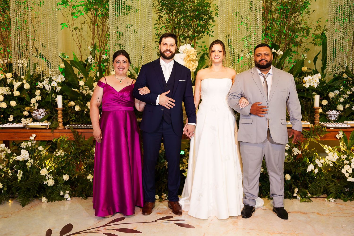 Wedding party of four posing at elegant floral ceremony backdrop with bride in white gown and guests in formal attire.