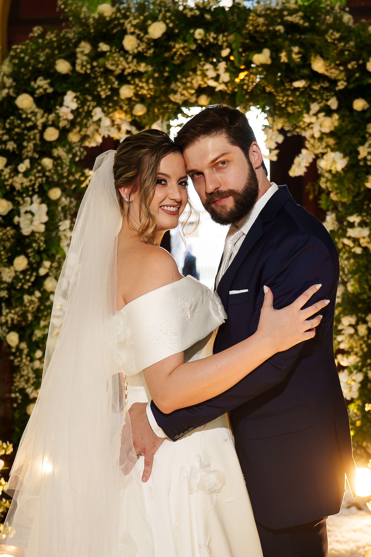 Newlywed couple embraces and smiles beneath an elegant white floral arch during their wedding celebration.