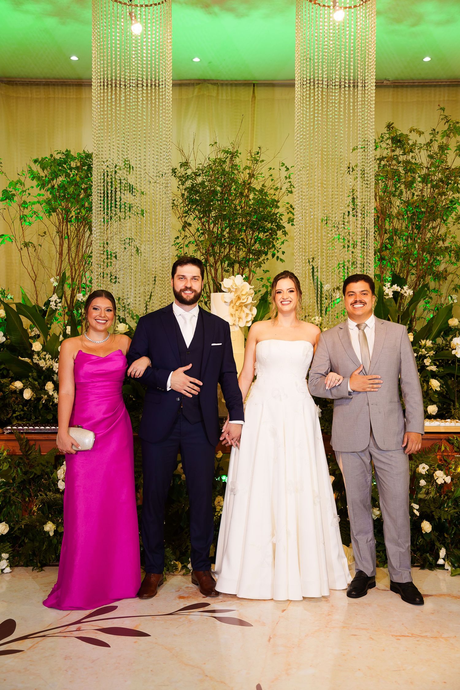 Bride and groom pose with two guests at elegant wedding reception with crystal chandeliers and lush greenery backdrop.