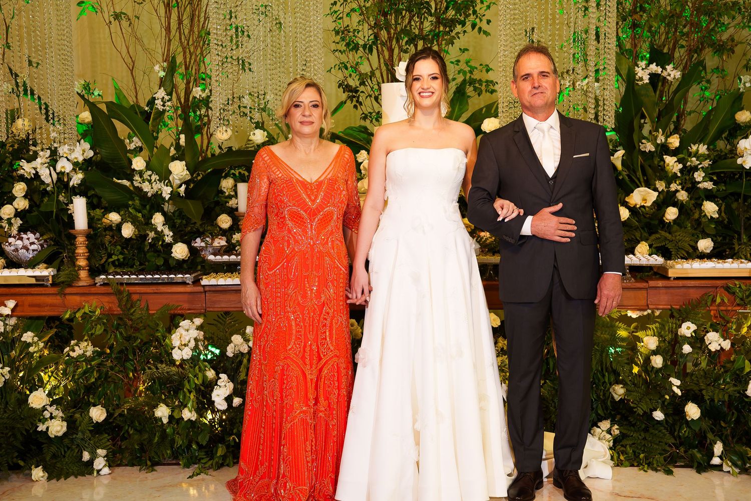 Bride in white gown poses with two guests at elegant wedding reception with floral backdrop.