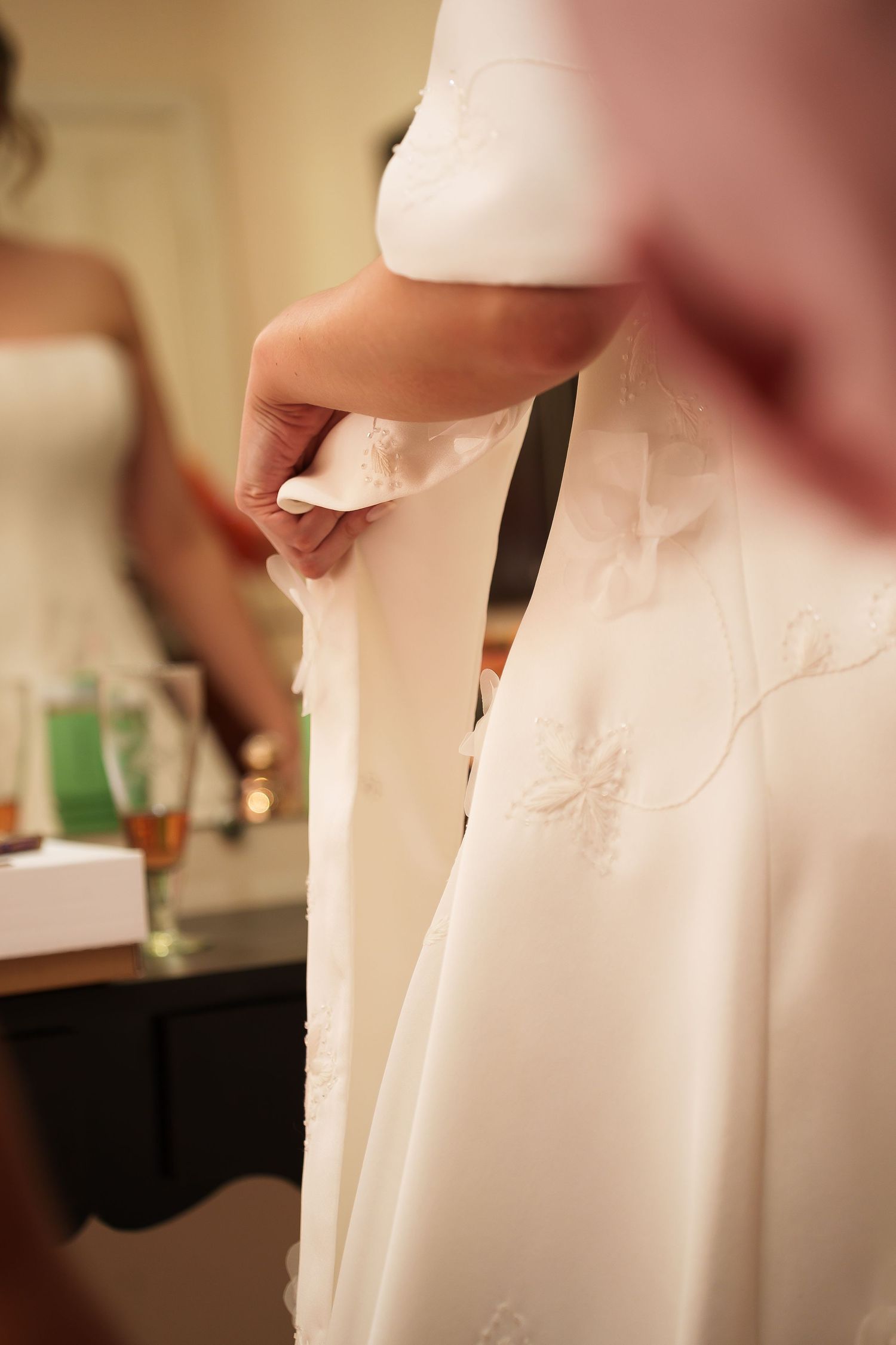Bride being helped into elegant white wedding gown with floral embroidery details, reflected in mirror.