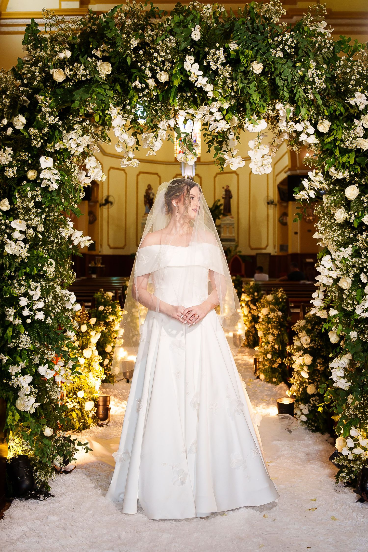 Bride in white off-shoulder gown and veil stands under floral arch with greenery and white flowers in church.