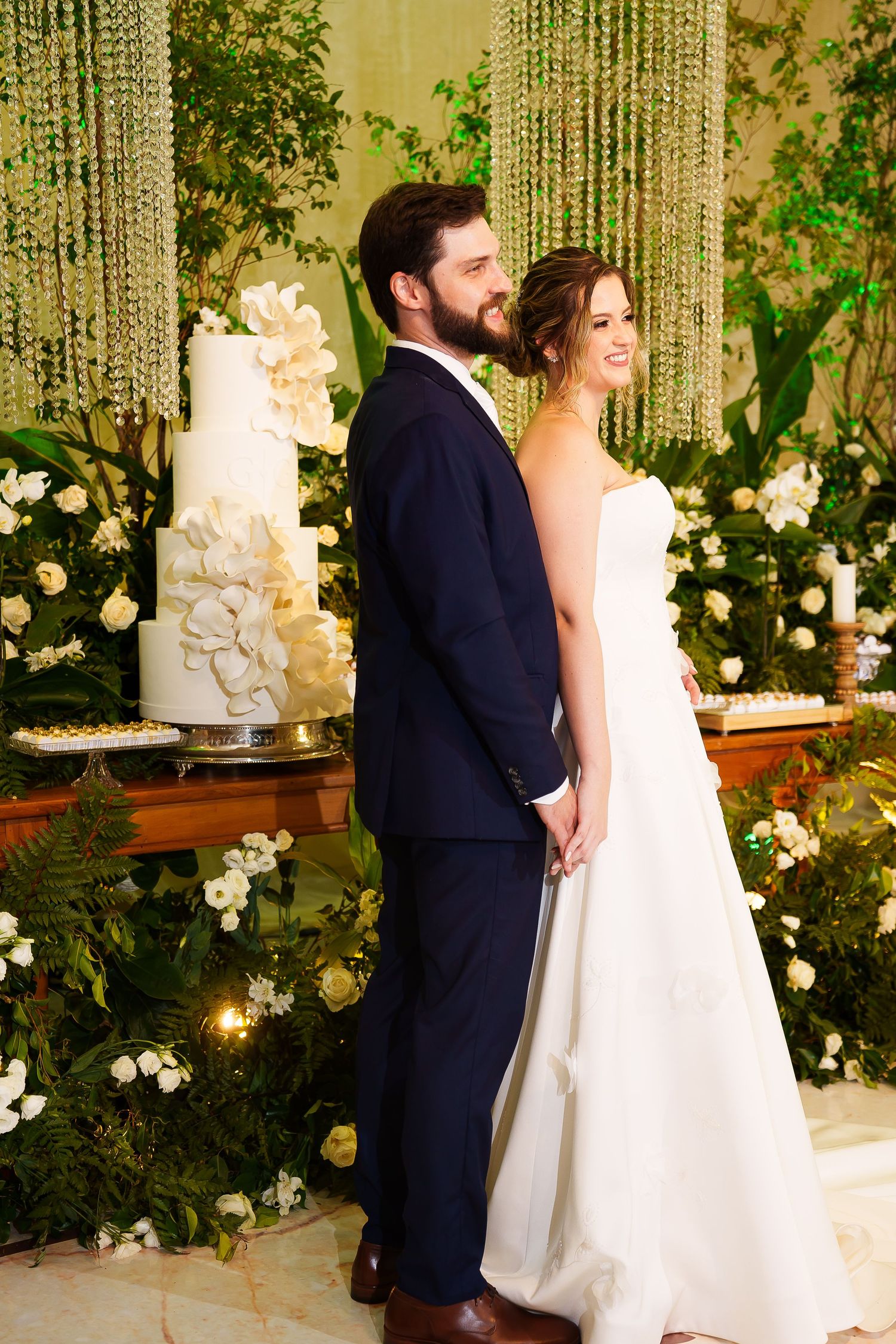 Bride and groom standing back-to-back at wedding reception with white floral cake and greenery backdrop.