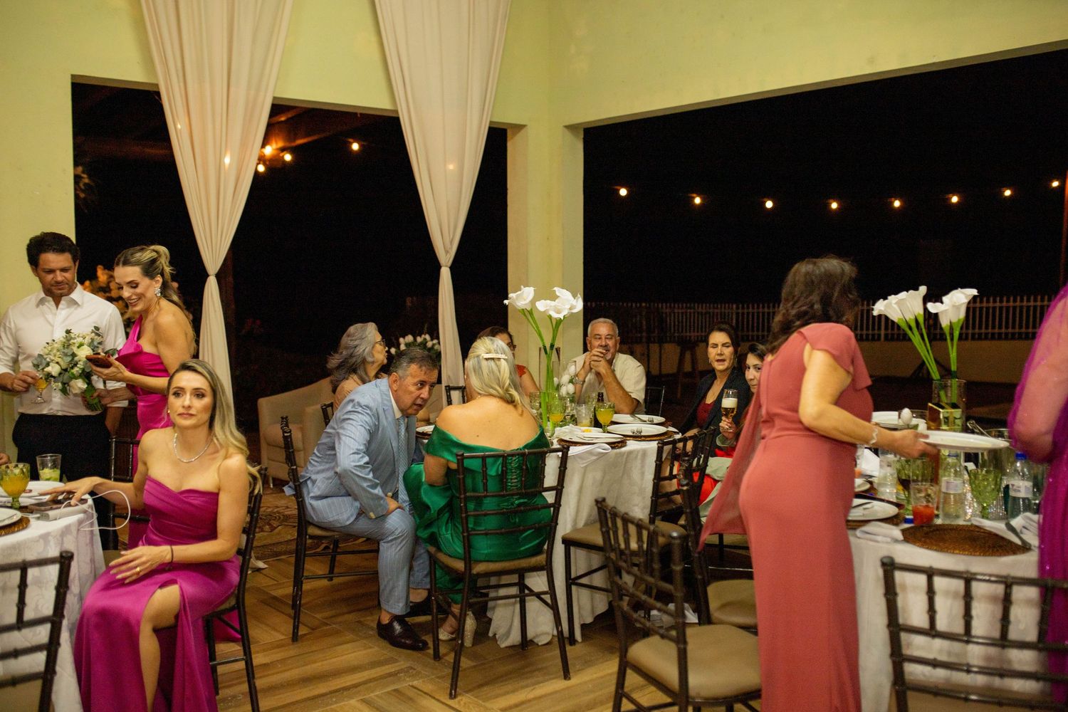 Elegant outdoor wedding reception with guests in formal attire seated at decorated tables under string lights.