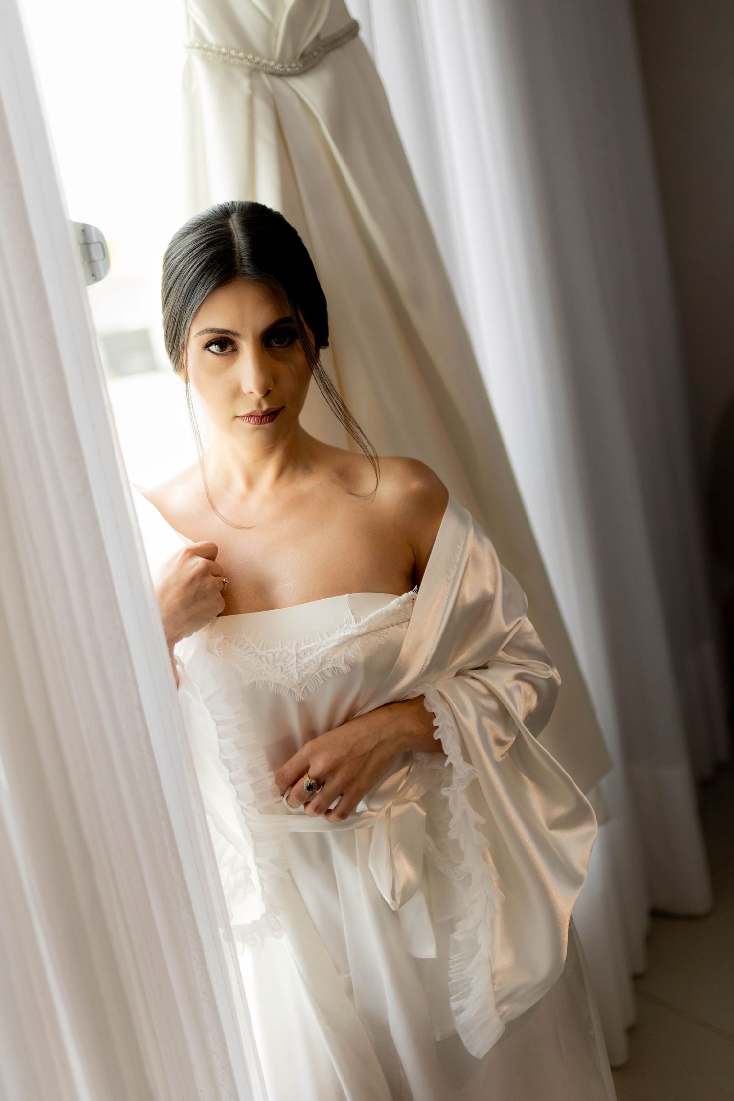 Bride in white lace corset and silk robe stands by curtains near her wedding dress in soft natural light.