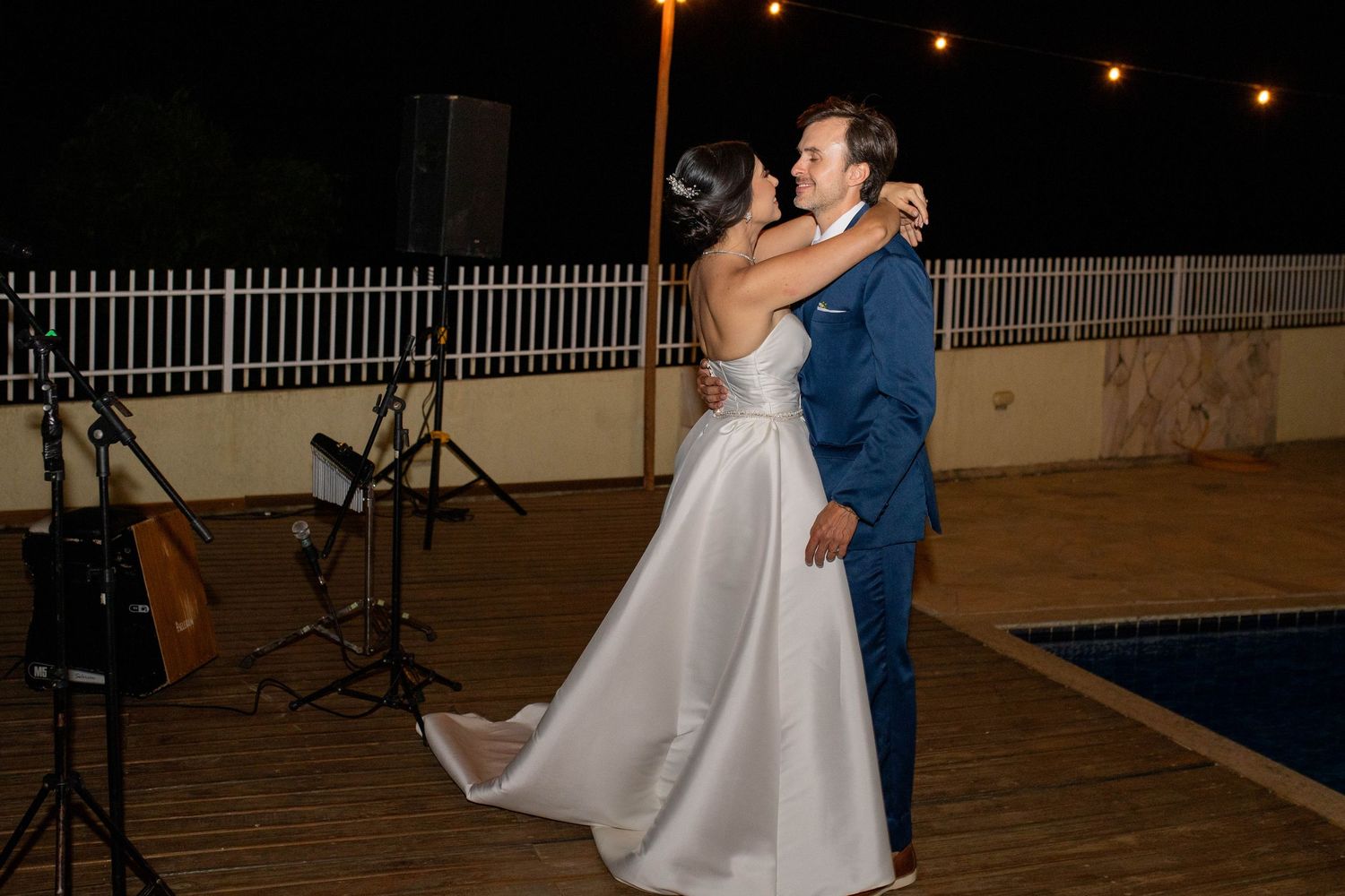 Bride and groom share their first dance outdoors at night near a pool with string lights and a live band.