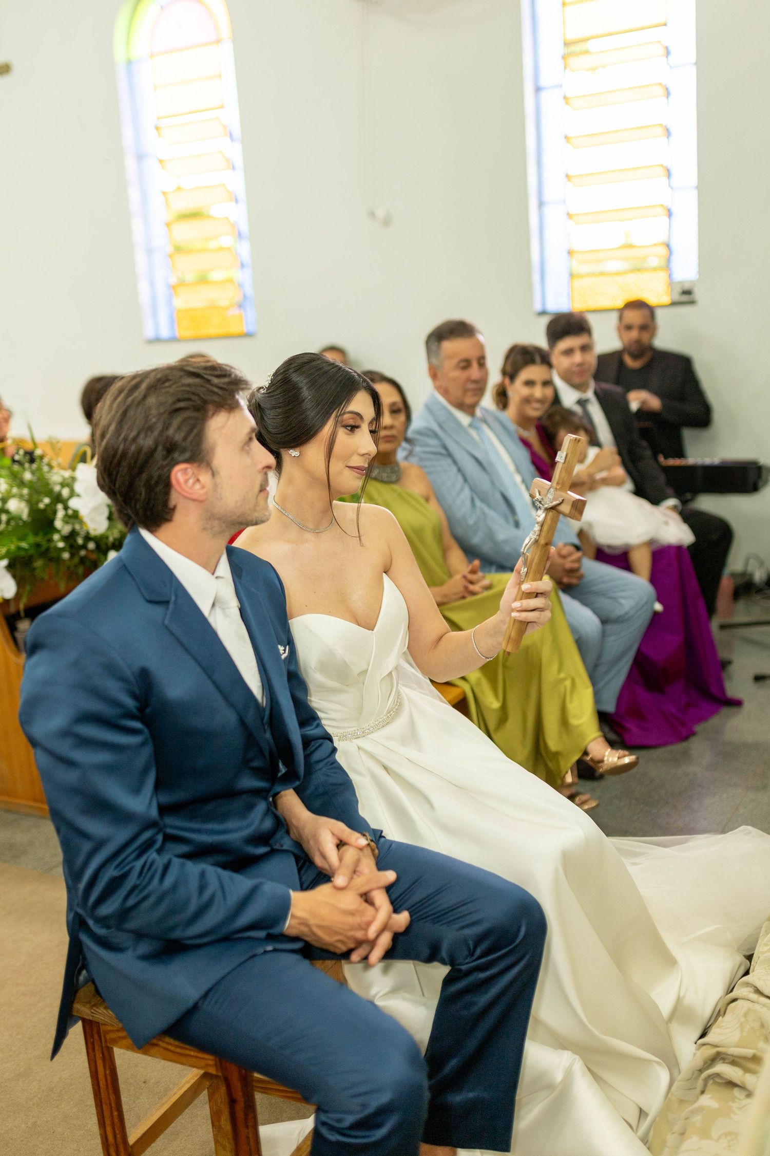 Bride and groom sit together during church wedding ceremony, bride holding a cross, guests seated in pews behind them.