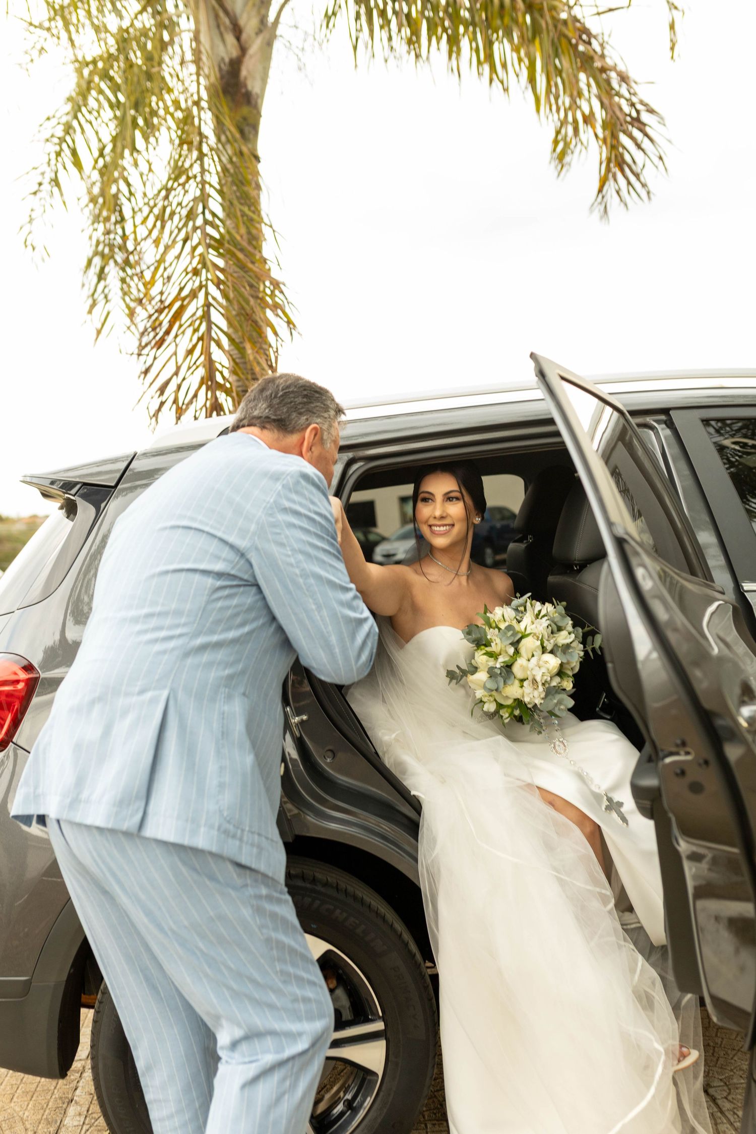 A smiling bride in a white gown holding a bouquet exits a car assisted by a man in a light blue suit near a palm tree.