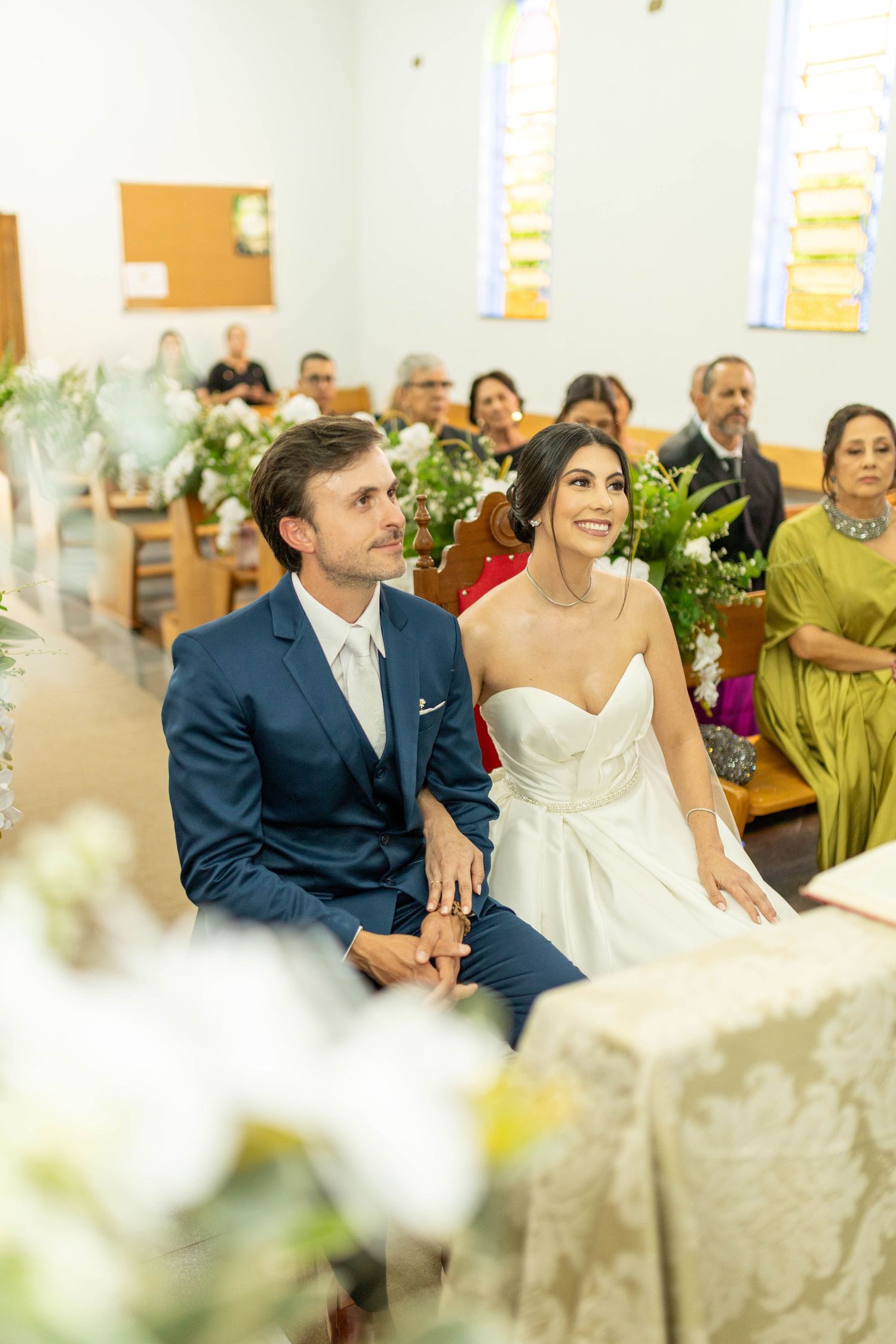 A bride and groom sit together at the altar during their church wedding ceremony, surrounded by guests and floral arrangements.