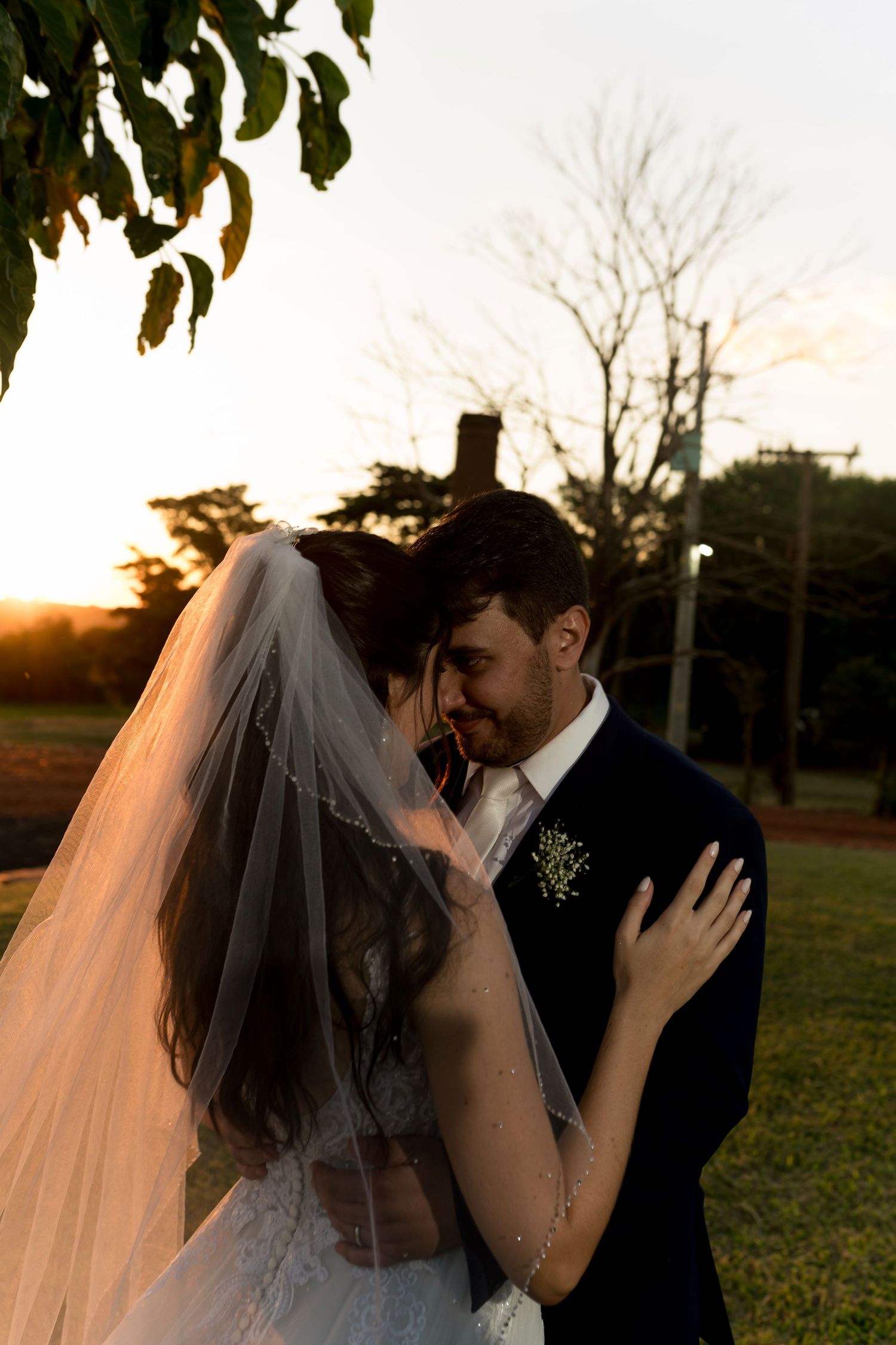 Bride and groom share a romantic sunset moment outdoors, with wedding veil flowing and groom wearing a boutonniere.