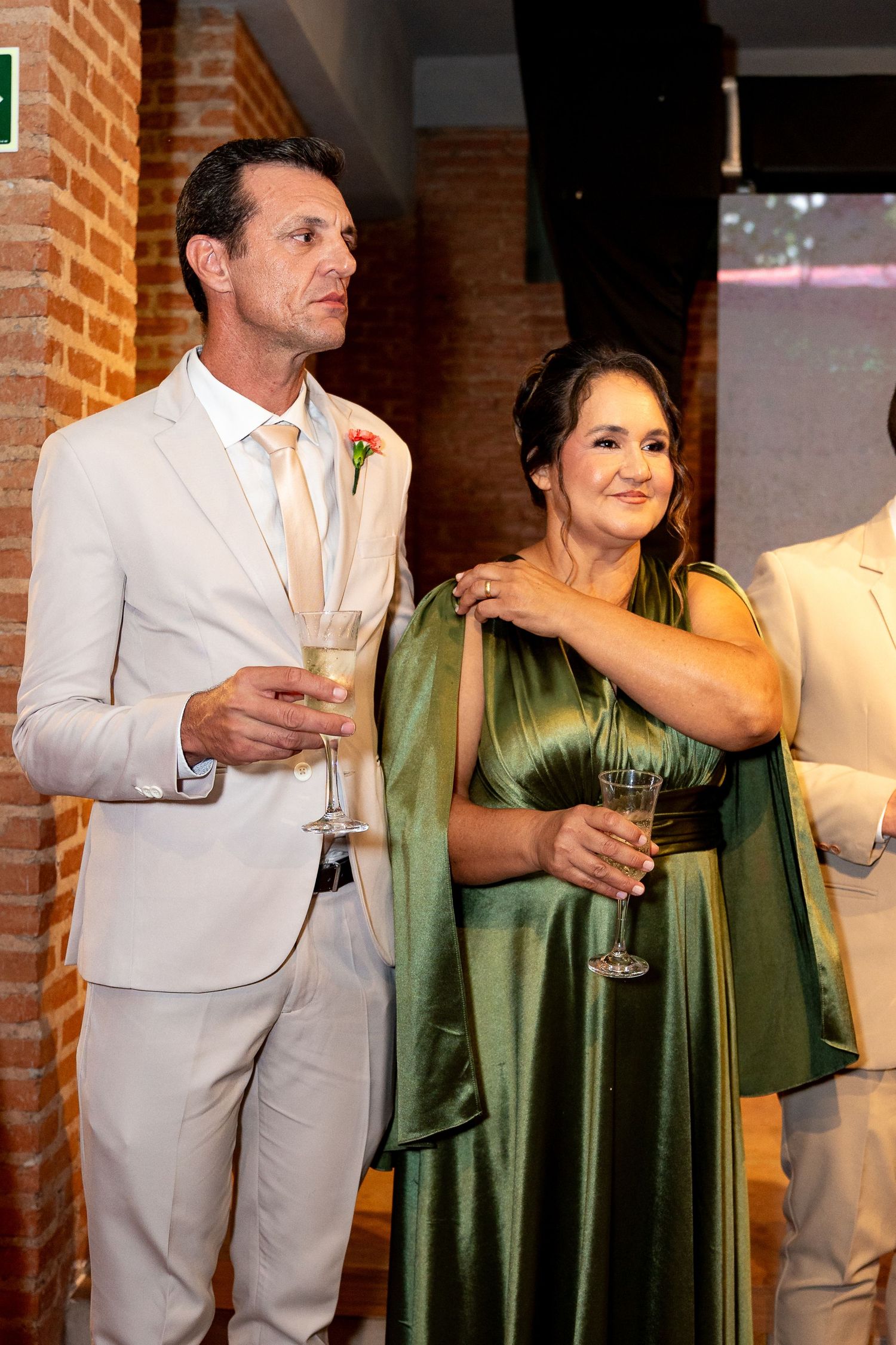 Elegant couple at wedding reception, man in white suit holding champagne, woman in green satin gown, brick wall backdrop.